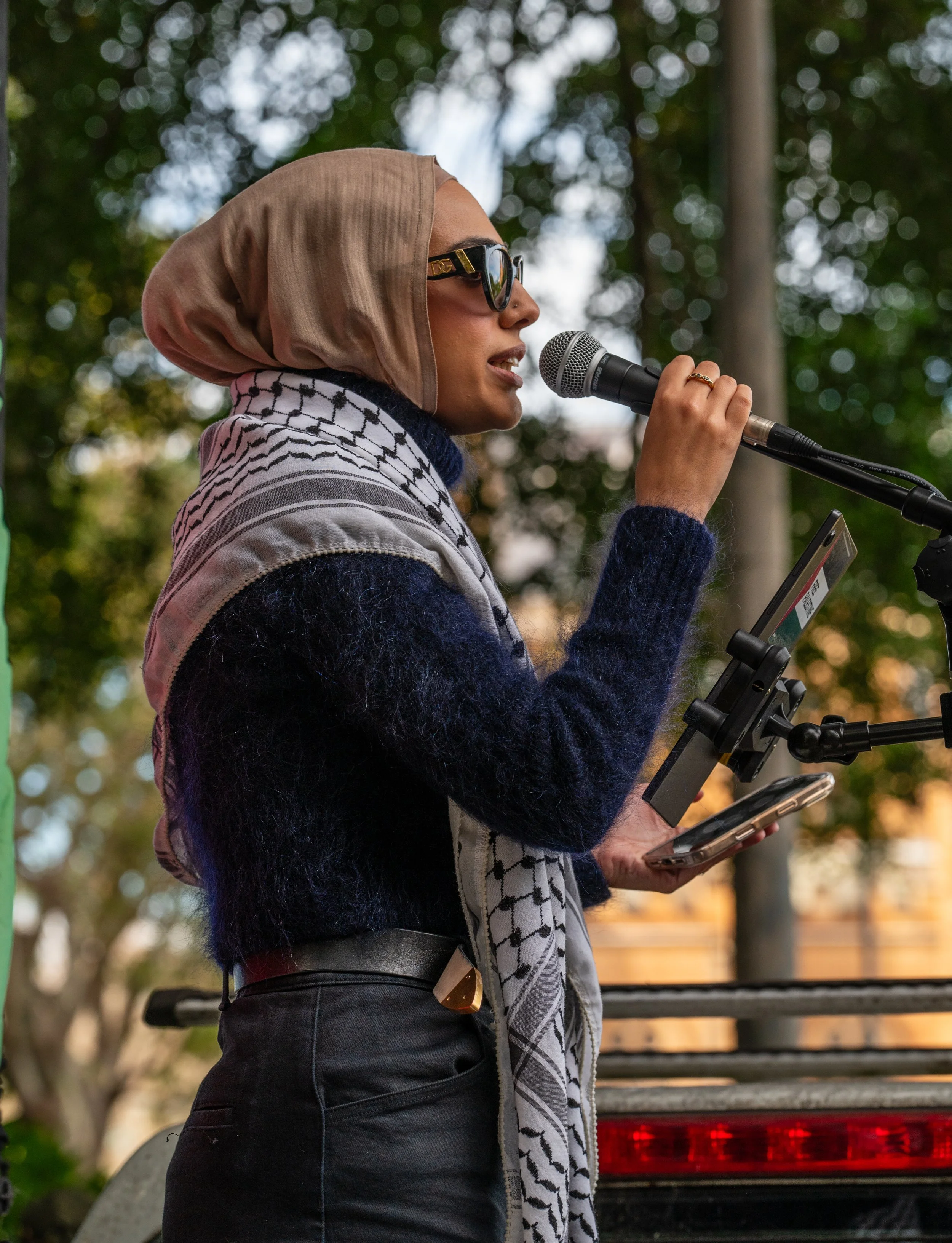 Woman wearing sunglasses, tan headscarf, black sweater, and hijab speaking into a microphone outdoors, holding a phone.