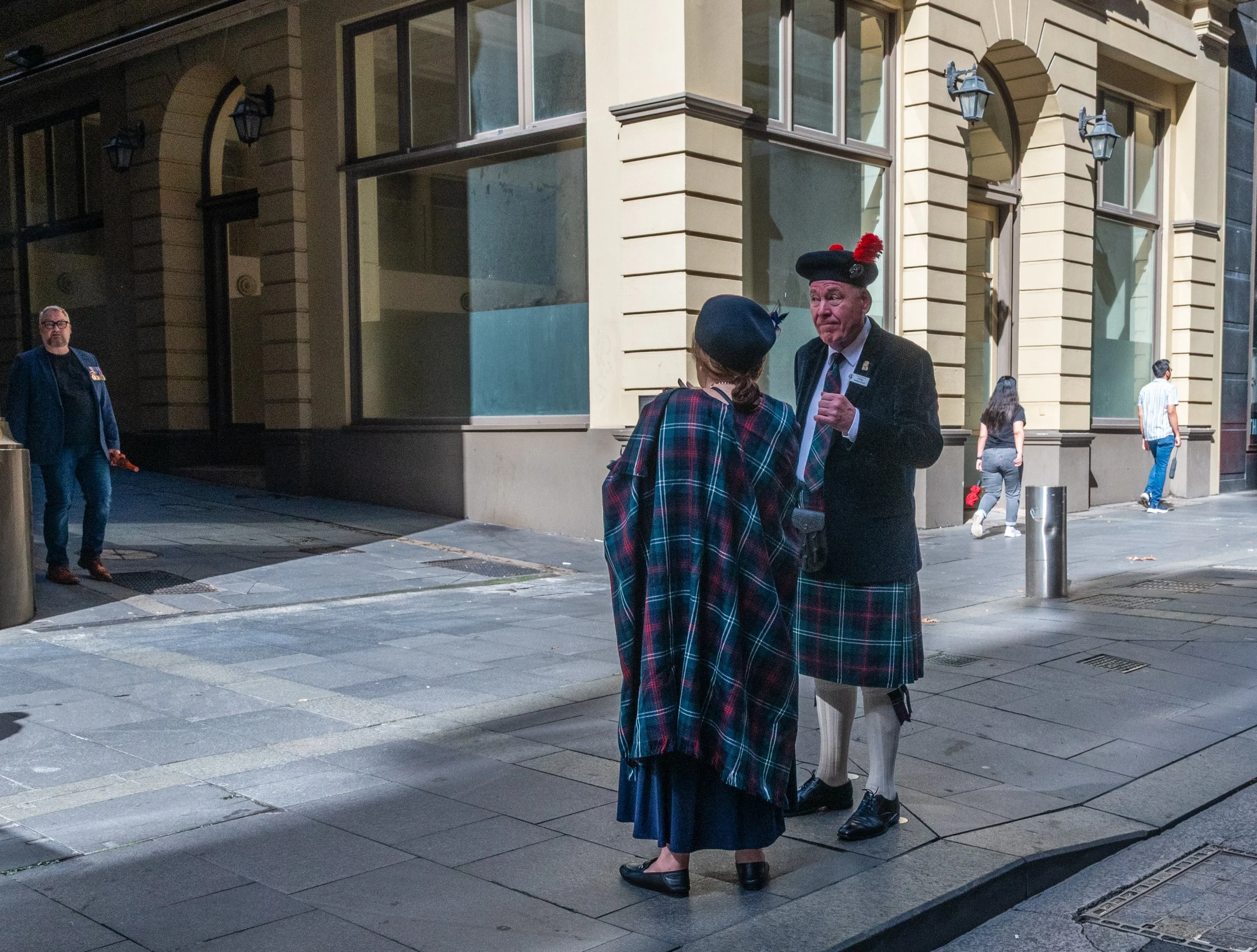 Two people dressed in traditional Scottish attire, a woman and a man, are talking on a city sidewalk at night. The man is wearing a black blazer, a plaid kilt, and a traditional Scottish hat with red pom-poms. The woman is wearing a plaid shawl and a dark skirt. They appear to be engaged in a conversation. In the background, a man in a blue jacket is walking, and two other pedestrians are walking on the sidewalk. The setting is an urban area with tall, light-colored buildings and street lamps.