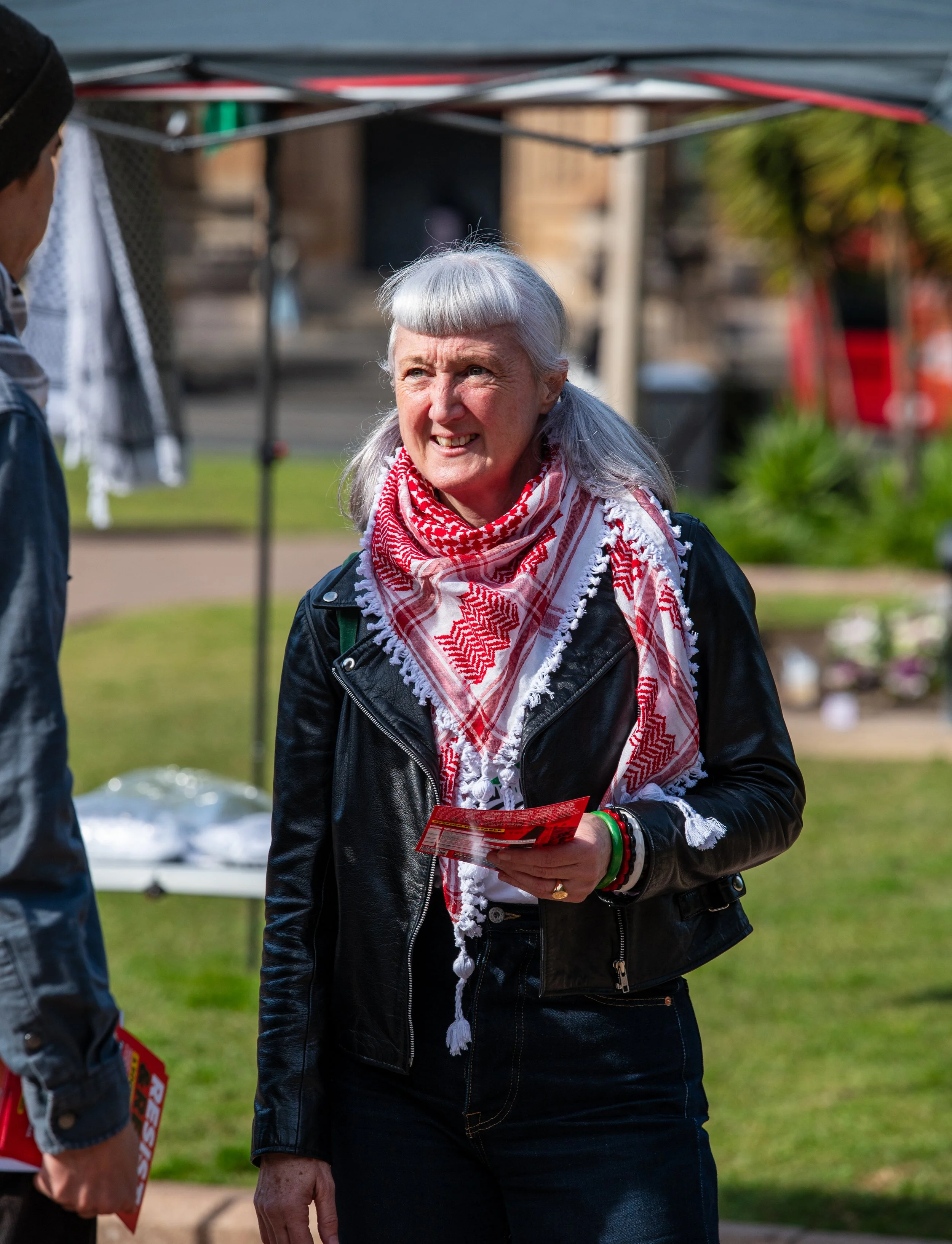 An older woman with gray hair styled in bangs and long hair, wearing a black leather jacket and a red and white keffiyeh scarf, is talking to a man at an outdoor event. She is holding a pamphlet and smiling.