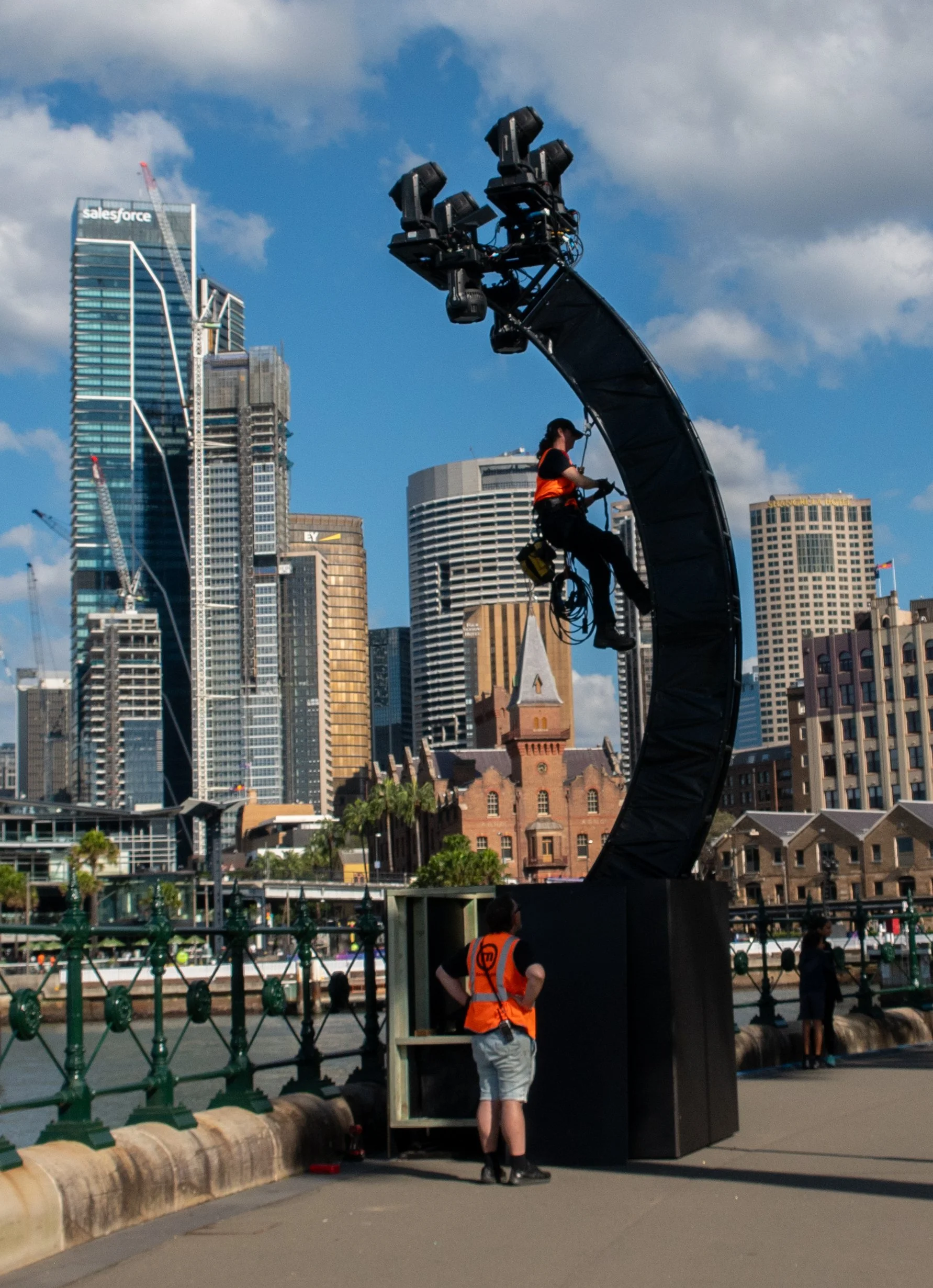A person working on a large stage light mounted on a curved structure, with an urban city skyline and skyscrapers in the background.
