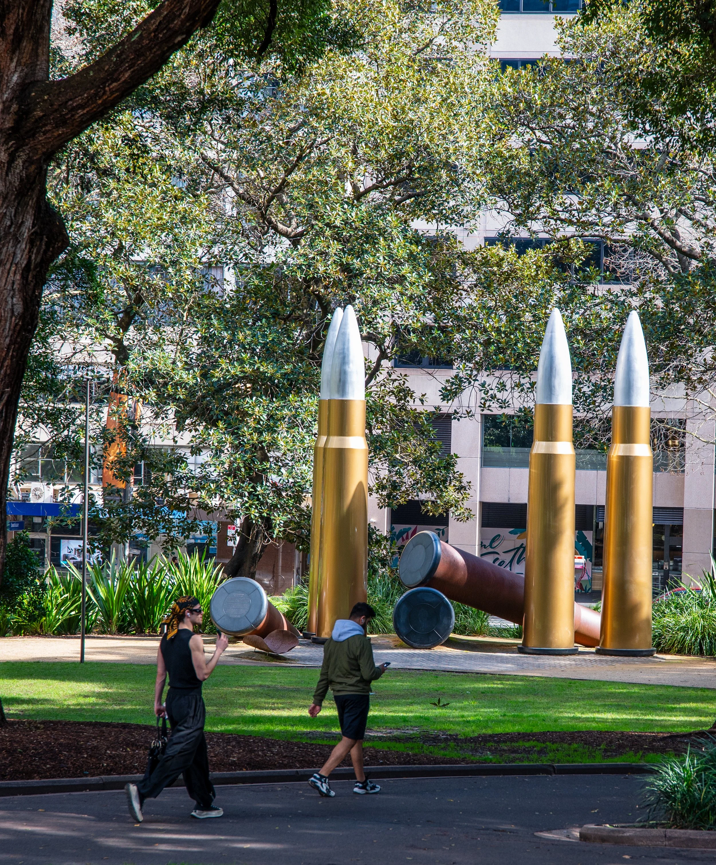 Public park with large sculpture resembling three bullets and two fallen shell casings, with people walking nearby.