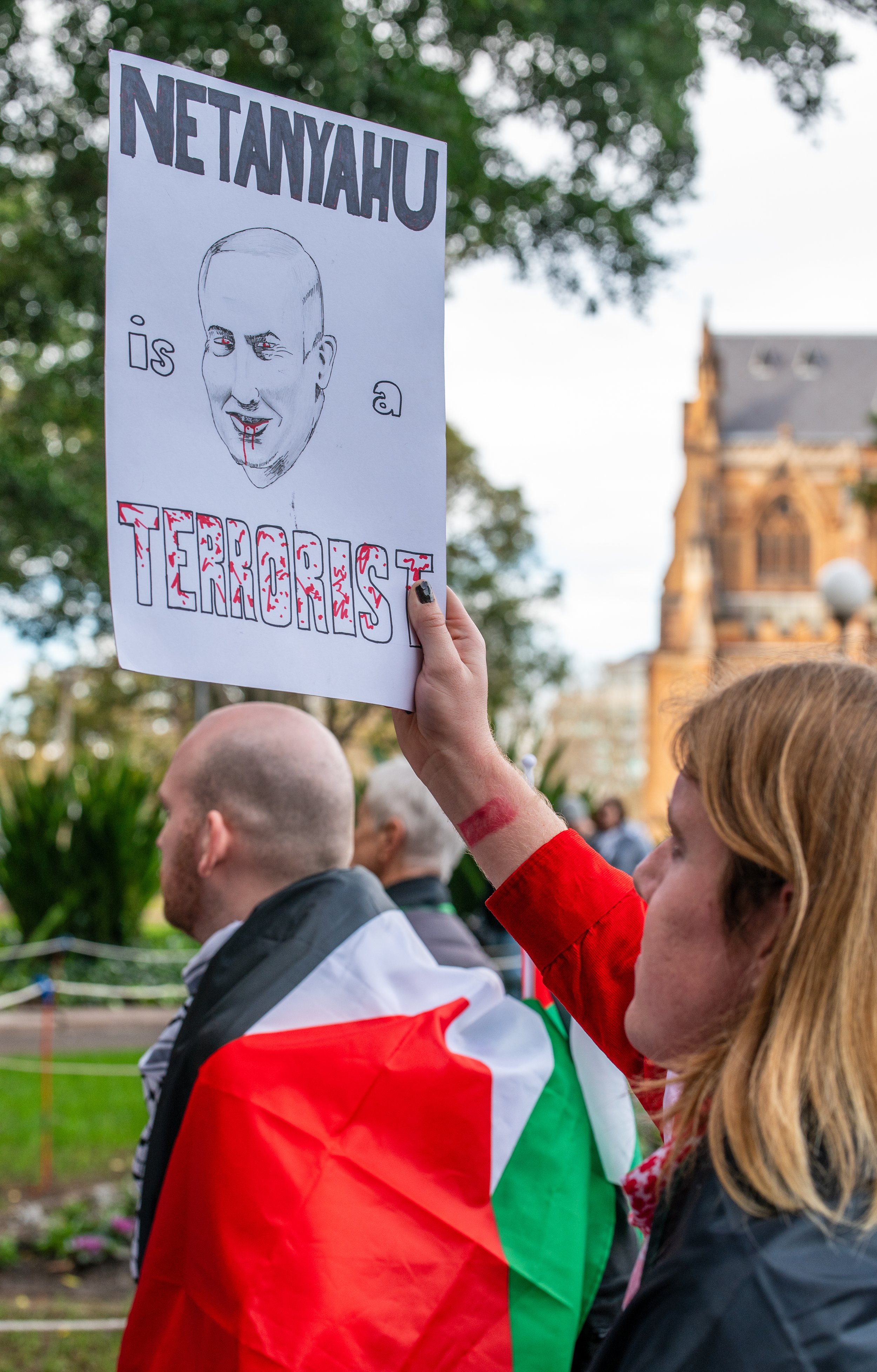 A woman holding a protest sign with a caricature of a man with blood on his face, with the text "NETANYAHU IS A TERRORIST". The woman has a red mark or tattoo on her arm, and a man draped in a Palestinian flag is visible nearby at an outdoor event.