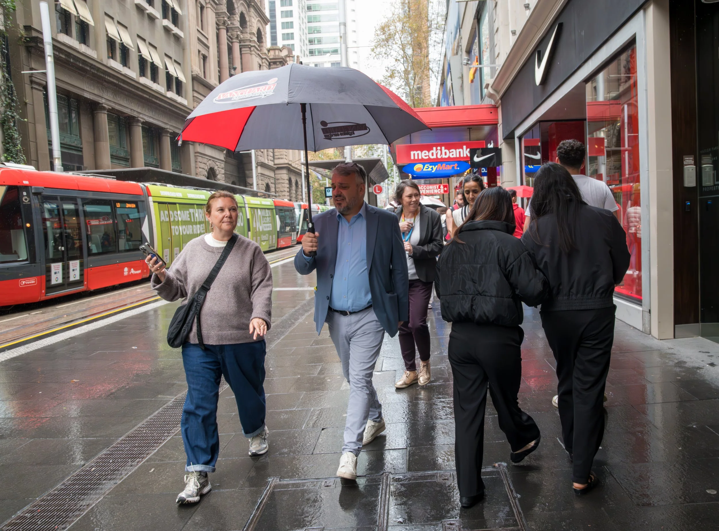 People walking on a rainy city sidewalk, some under an umbrella, with a red tram, buildings, and storefronts in the background.