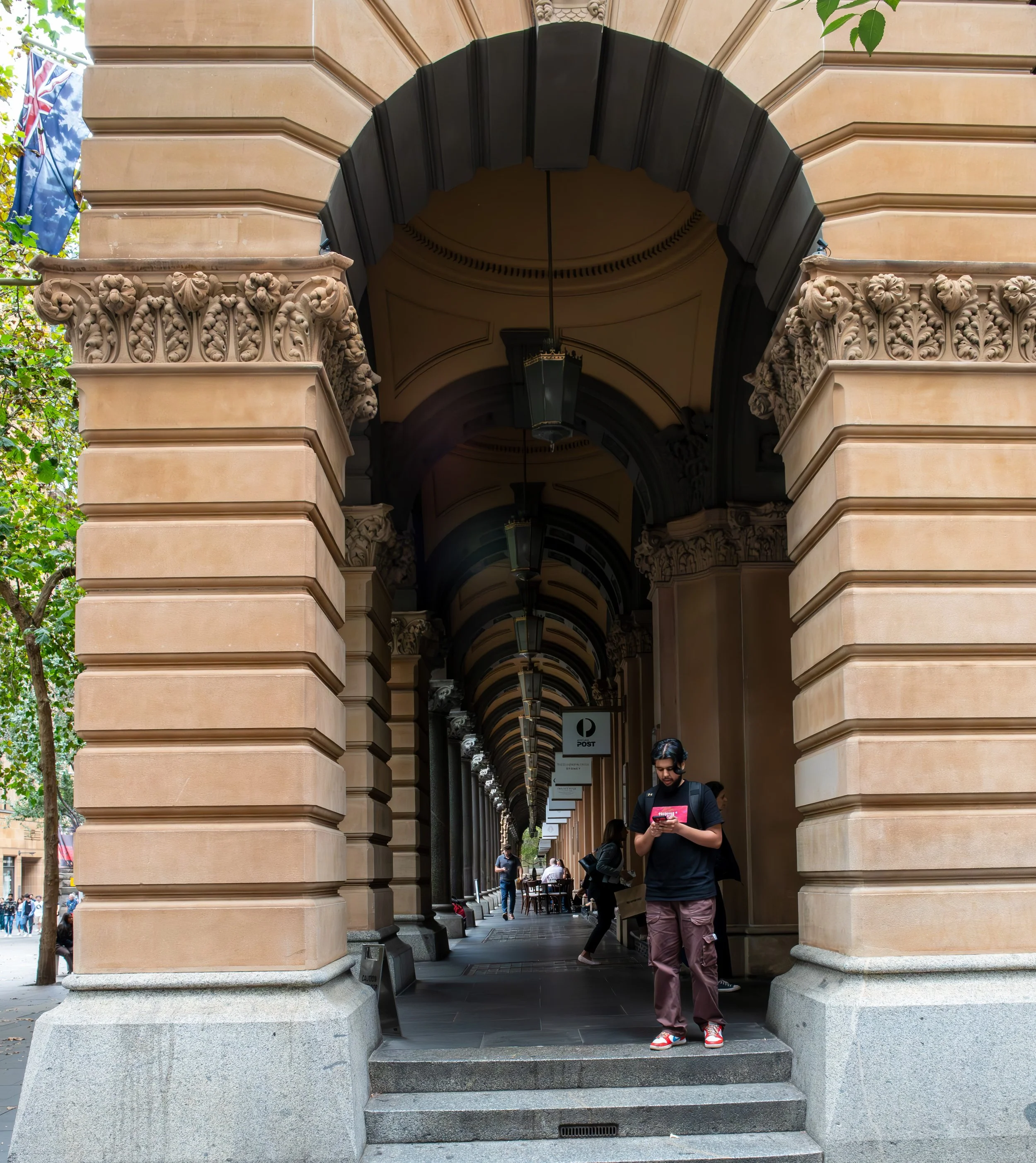 A man standing under a historic arched walkway, looking at his phone with other people walking and sitting in the background.