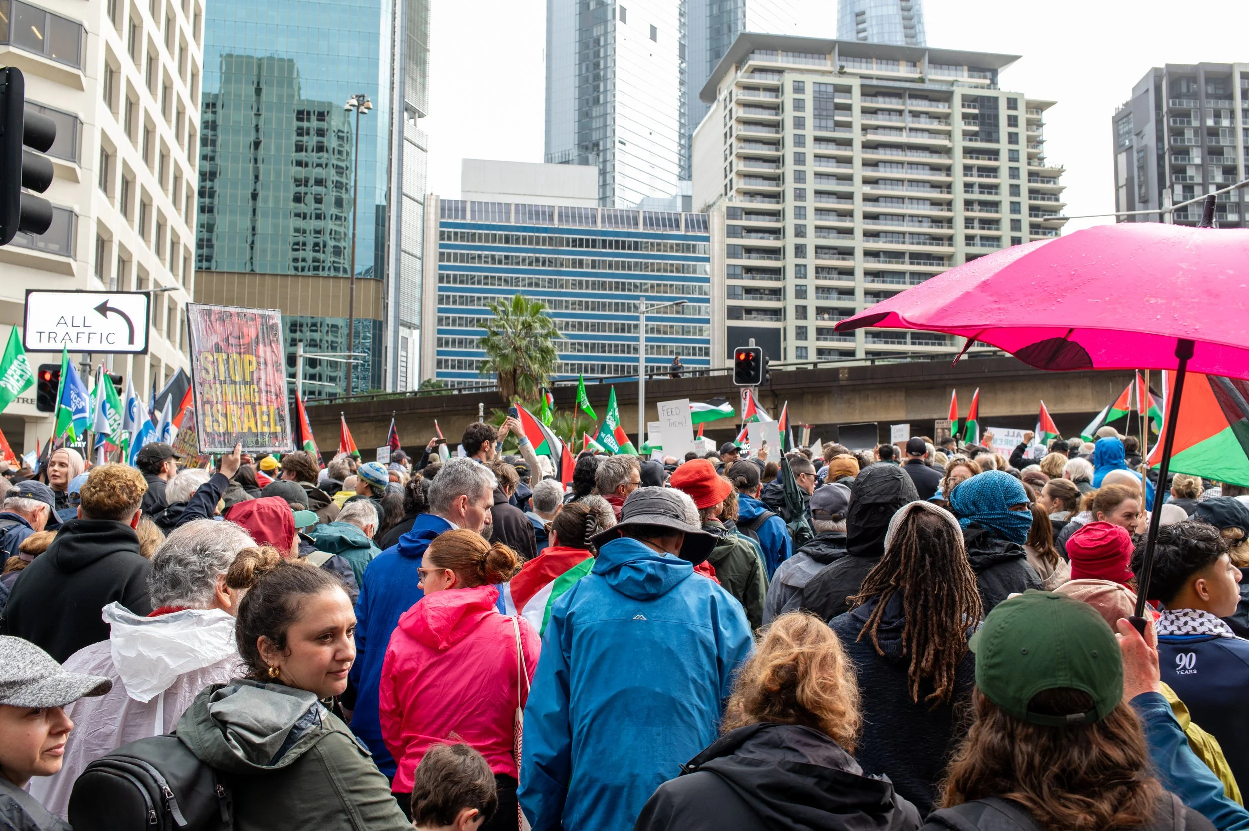 Crowd of people protesting in an urban area with skyscrapers in the background, holding signs and flags, some under a pink umbrella.