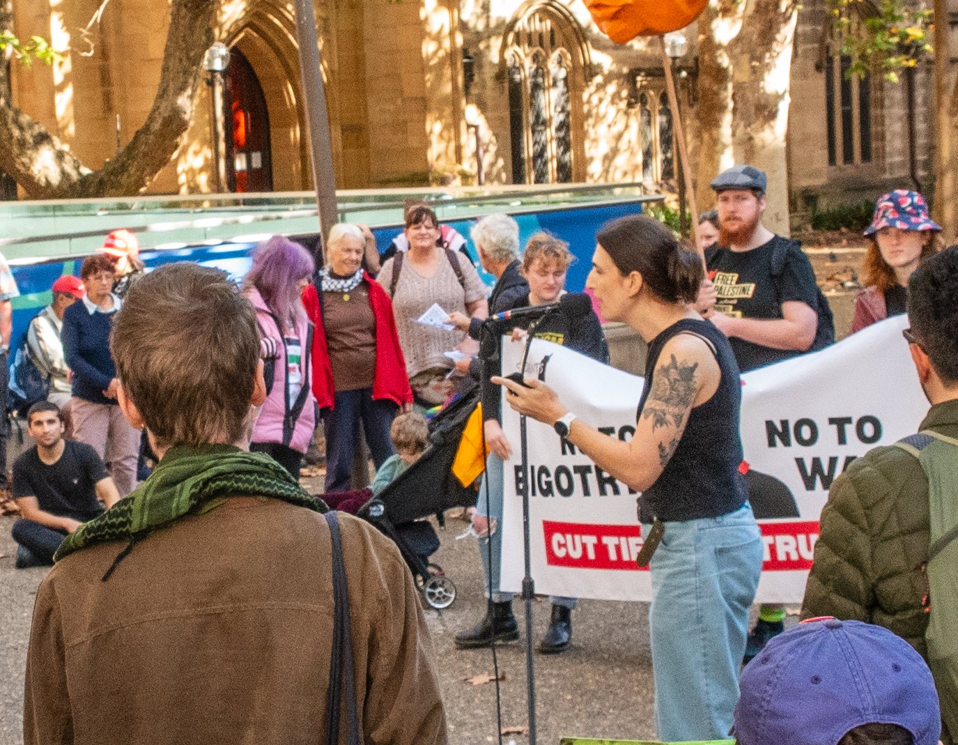A woman with tattoos on her arm speaking into a microphone at a protest. Behind her, a group of diverse people hold a banner with slogans such as "NO TO WAR" and "CUT TIES." The protest takes place outdoors, near an old building with trees providing shade.