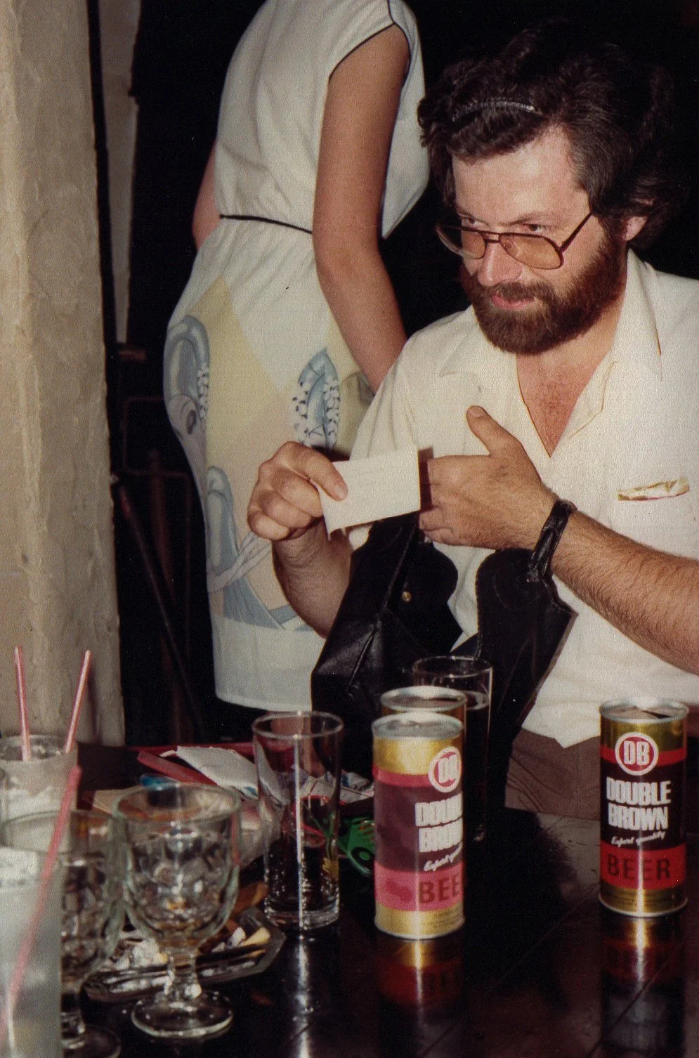A man with glasses and a beard is sitting at a table, looking at a piece of paper. There are cans of Double Brown beer and glasses with drinks on the table. A woman, partially visible, is standing nearby with her back to the camera, wearing a dress with artwork on the back.