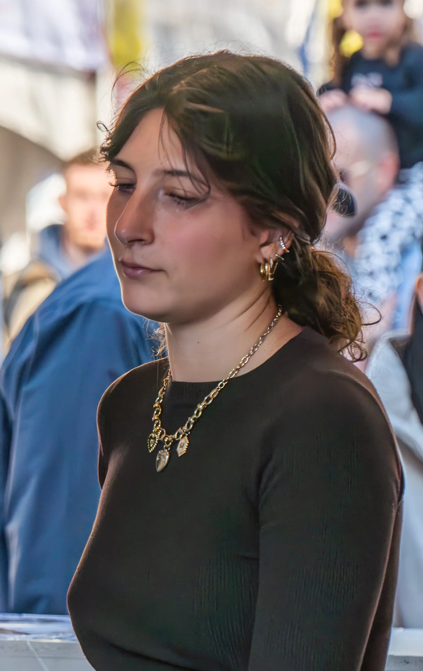 A woman with dark hair and gold jewelry, wearing a black top, has her eyes closed and appears to be in a contemplative or peaceful moment, with blurred people in the background.