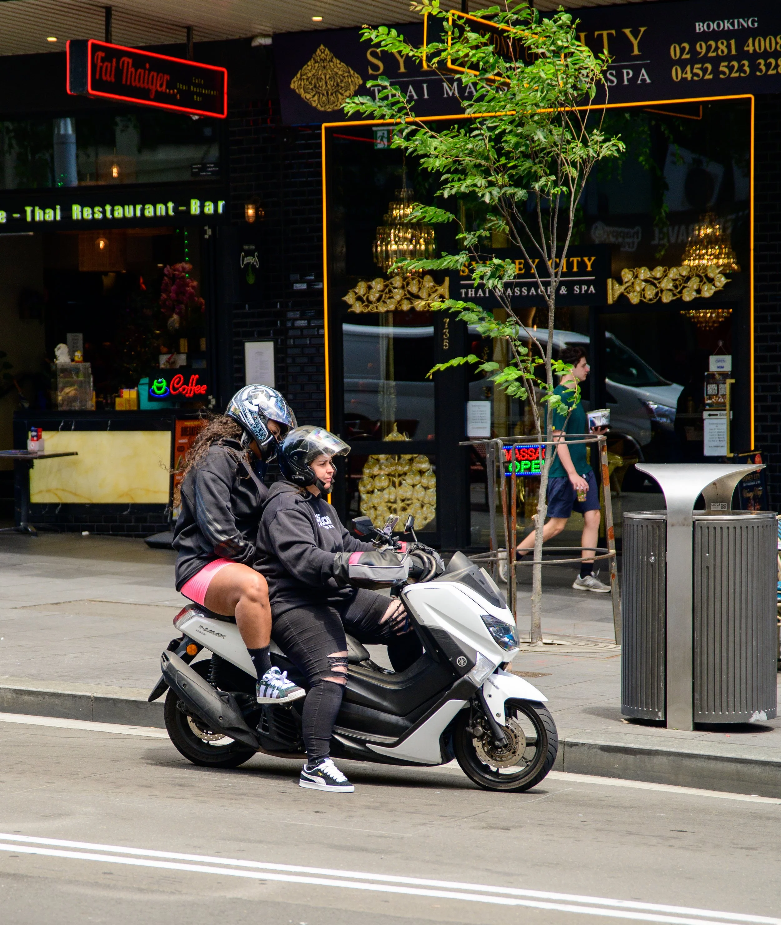Two people riding a scooter on the street in front of a Thai massage and spa shop. The rider in front is a young woman wearing black ripped jeans and a black hoodie, the passenger is wearing pink shorts.