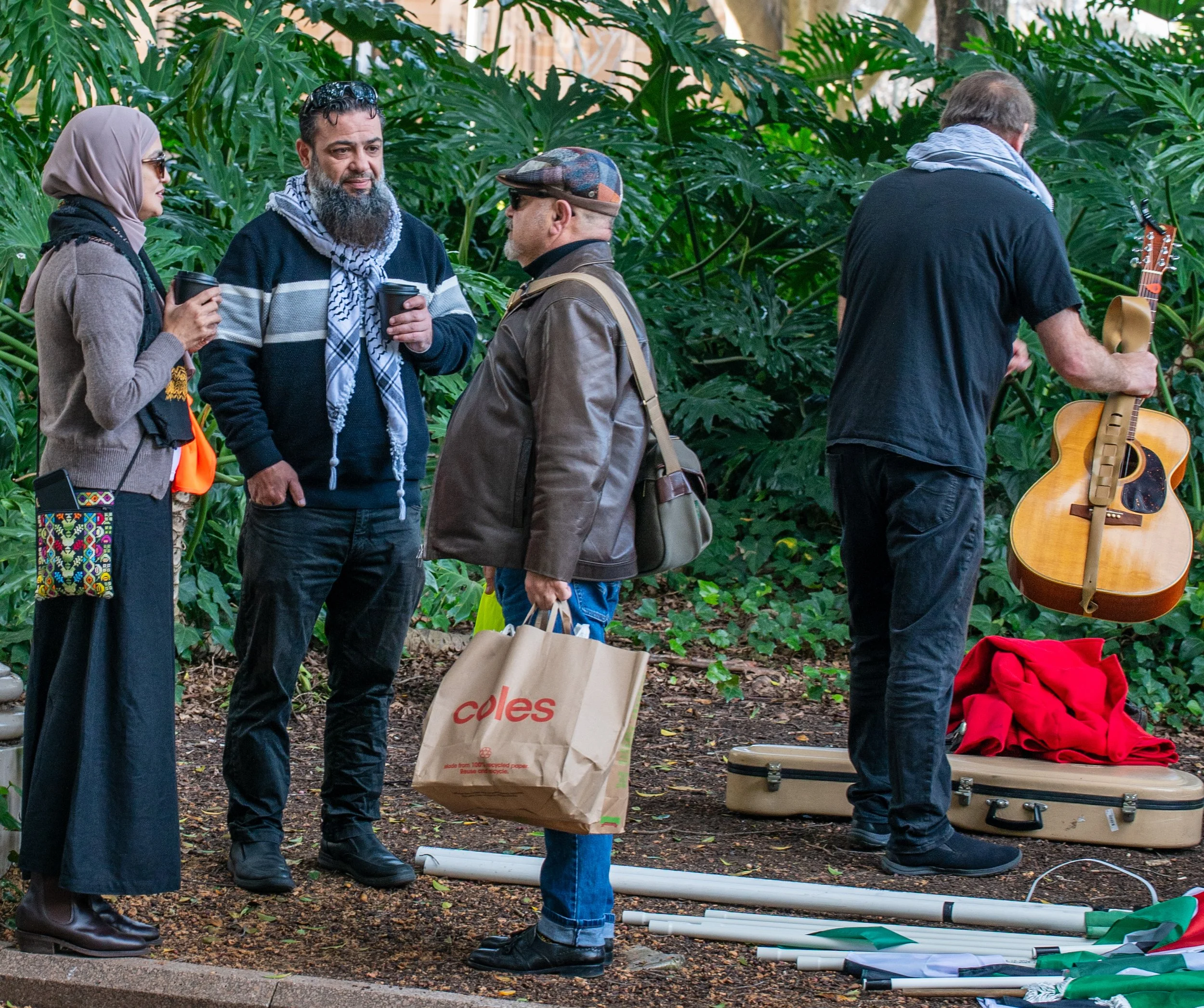 Four people in a greenhouse with plants, two of them talking while holding coffee cups, one adjusting a guitar, and another holding a shopping bag, with musical equipment and red clothing nearby.