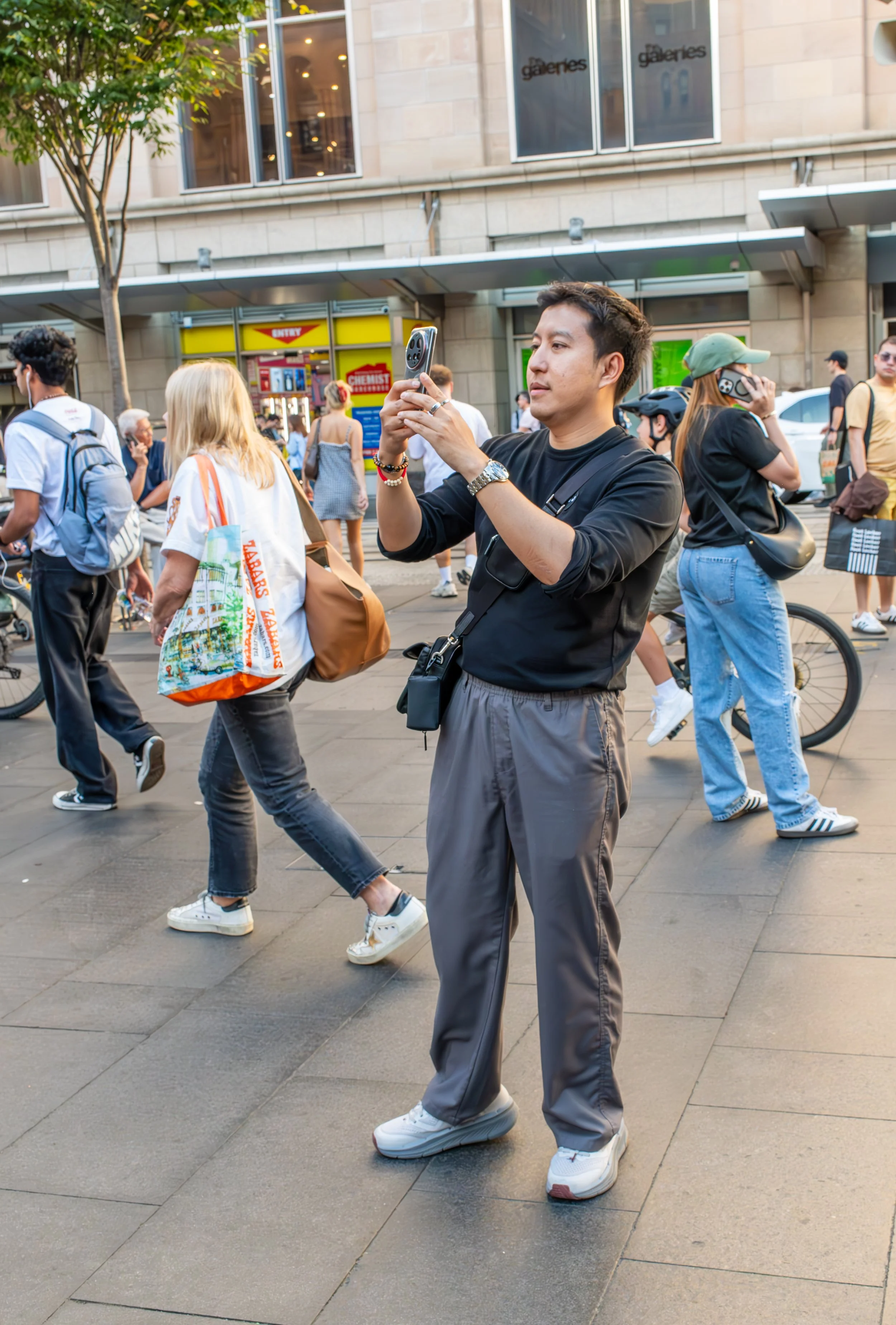A man in a black shirt and gray pants is taking a photo with his phone on a busy city street. Several pedestrians, some with backpacks and bicycles, walk past him, and storefronts are visible in the background.