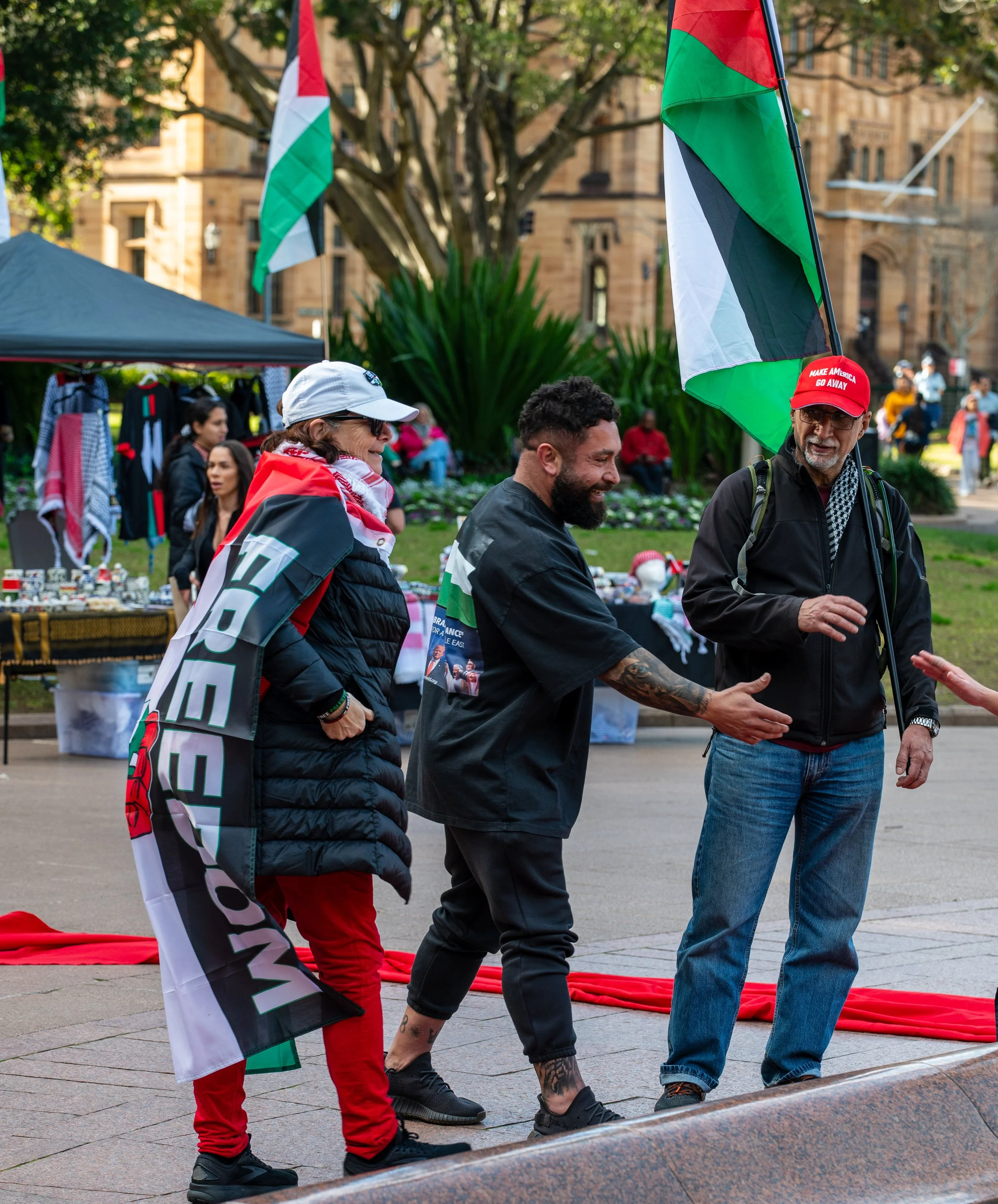Three people engaged in conversation at a protest or rally, with Palestinian flags and merchandise visible in the background.