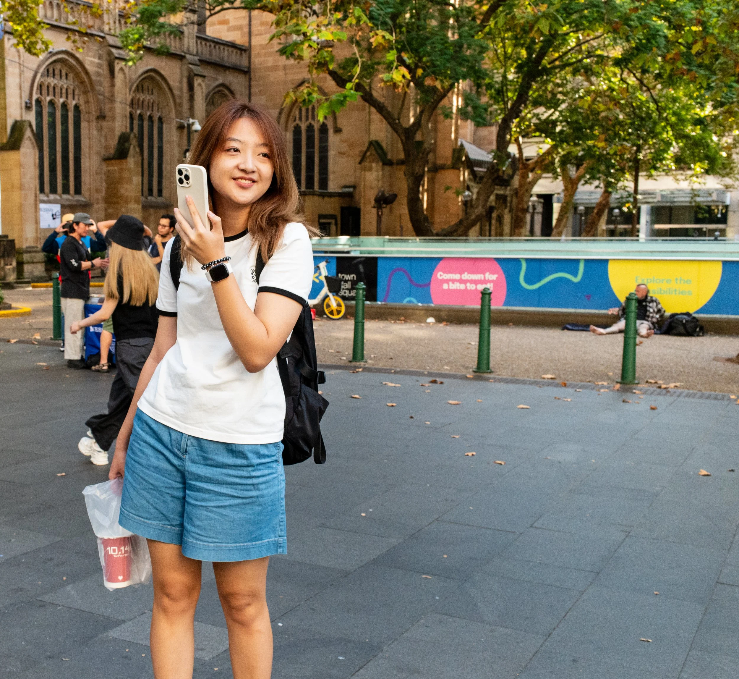 A young woman with shoulder-length brown hair, wearing a white T-shirt with black trim and a denim skirt, holding a phone in her right hand and a plastic bag in her left, standing on a city street. There are people and a historic stone building with arched windows in the background, along with a green tree and a colorful advertising board.