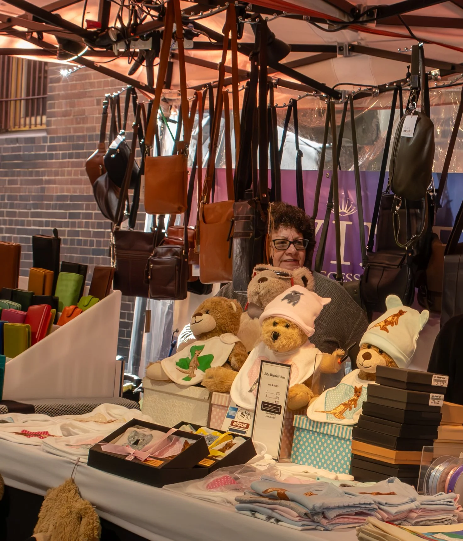 A woman selling plush teddy bears, purses, and other accessories at an outdoor market stall with a brick wall in the background.