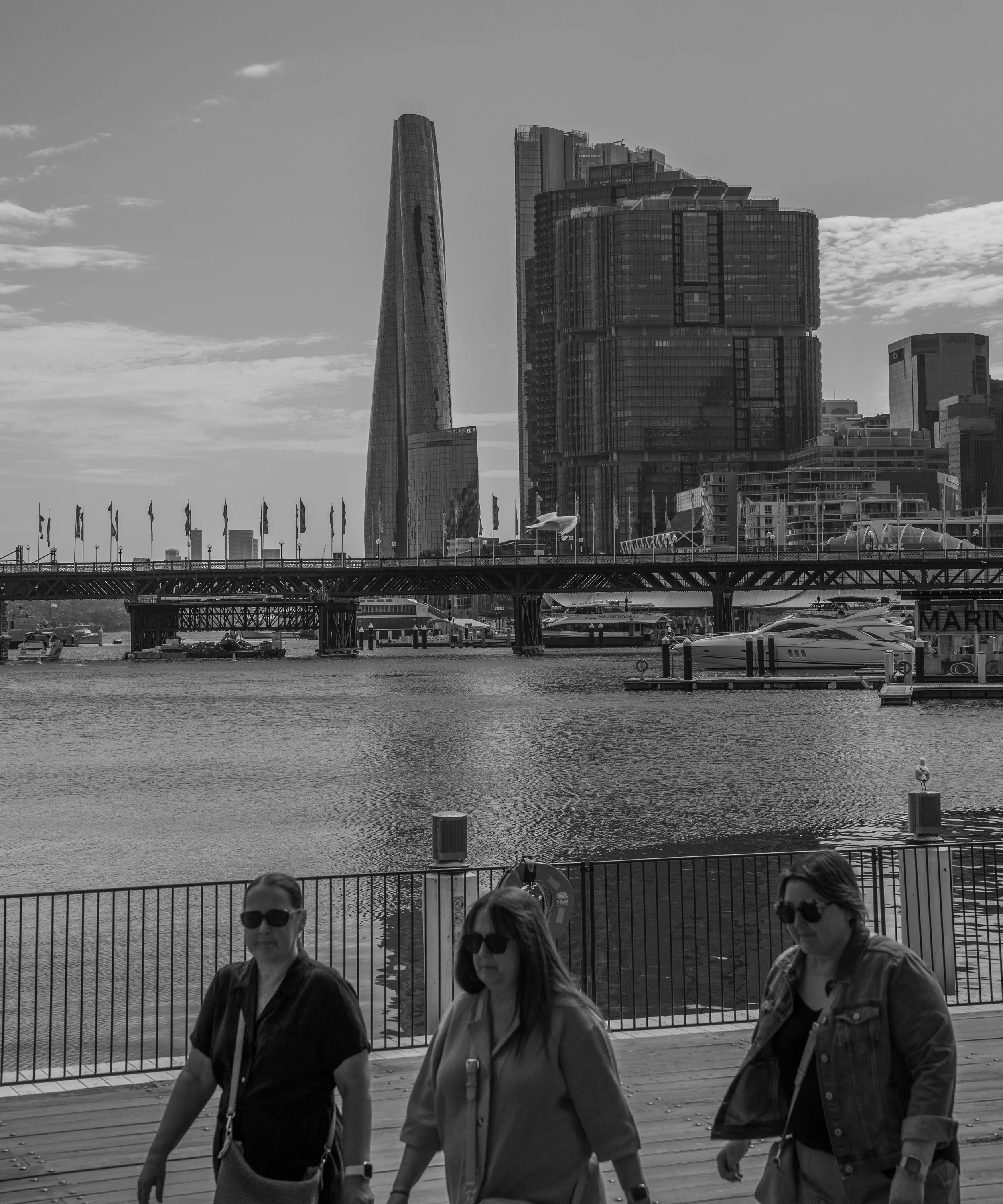 Three women walk along a waterfront promenade with the city skyline and river in the background.