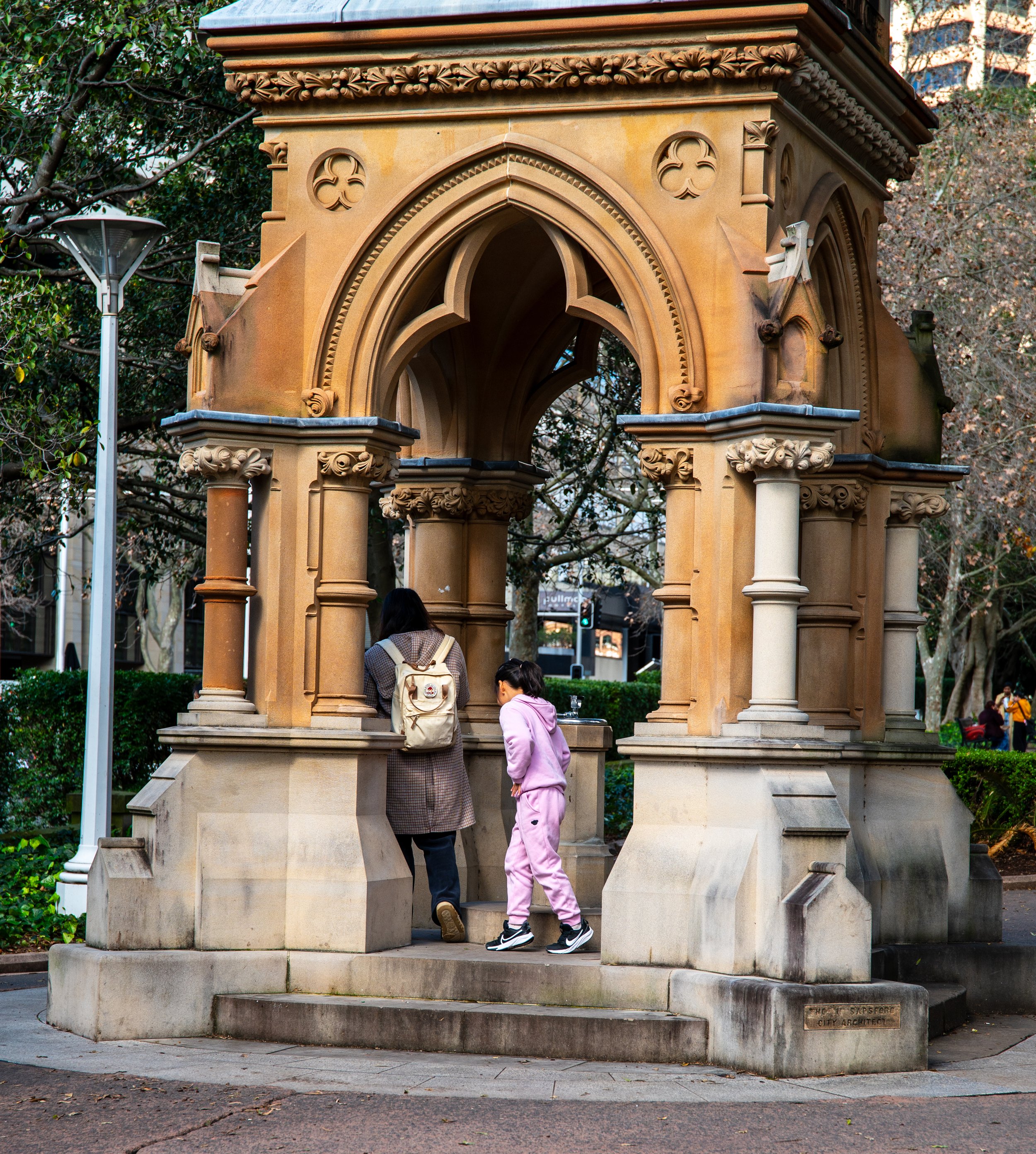 A historic stone fountain with gothic architecture in a park, featuring two people, a woman with a backpack and a child in pink clothing, near the fountain.