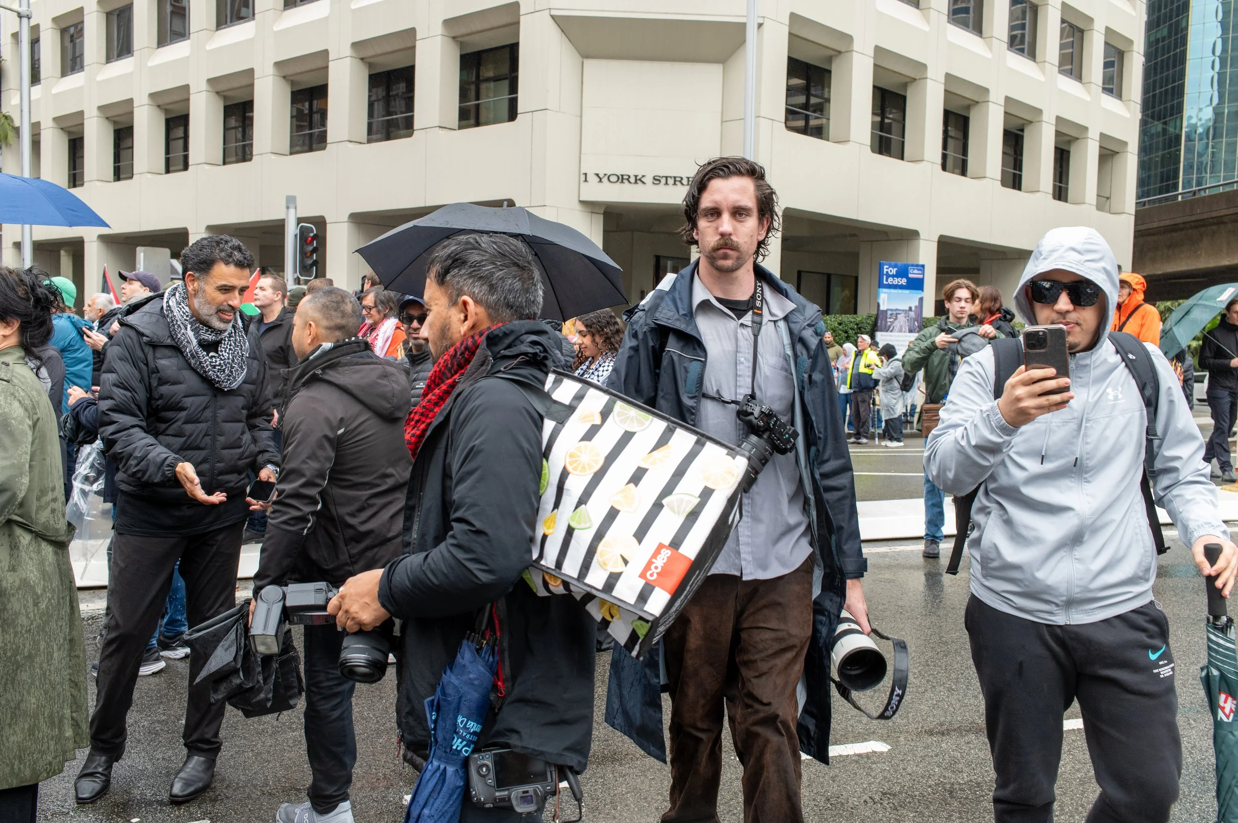 A group of people, some holding umbrellas, gathered on a wet city street in front of a modern building, many with cameras and phones, during rainy weather.
