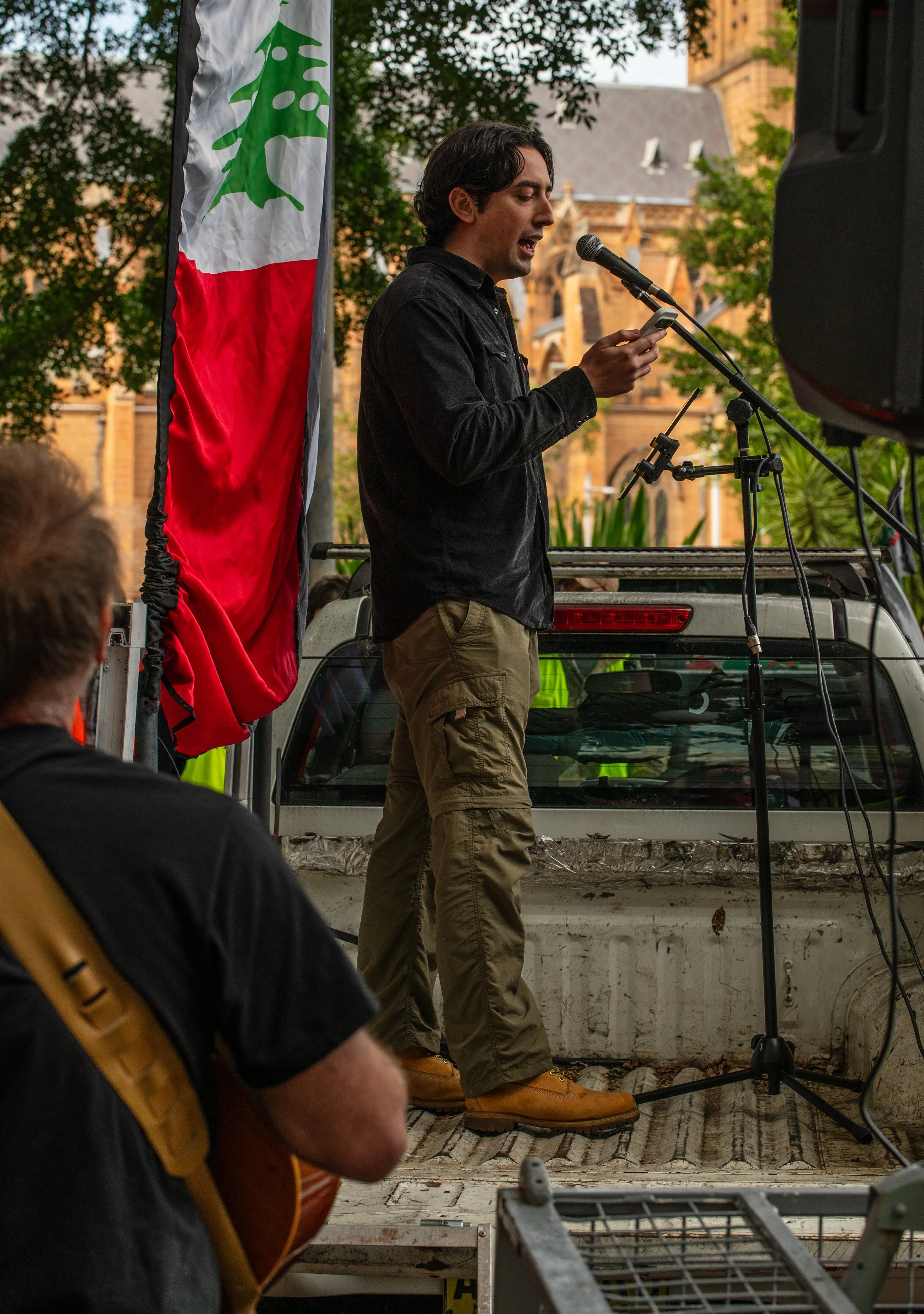A man with dark hair, dressed in a black shirt and tan cargo pants, stands on the back of a truck, speaking into a microphone while looking at his phone. A flag with green, white, and red stripes and a green cedar tree emblem is beside him. In the foreground, a person with a guitar, wearing a black shirt, is seated and facing the speaker. The setting appears to be an outdoor event in front of a historic building and trees.