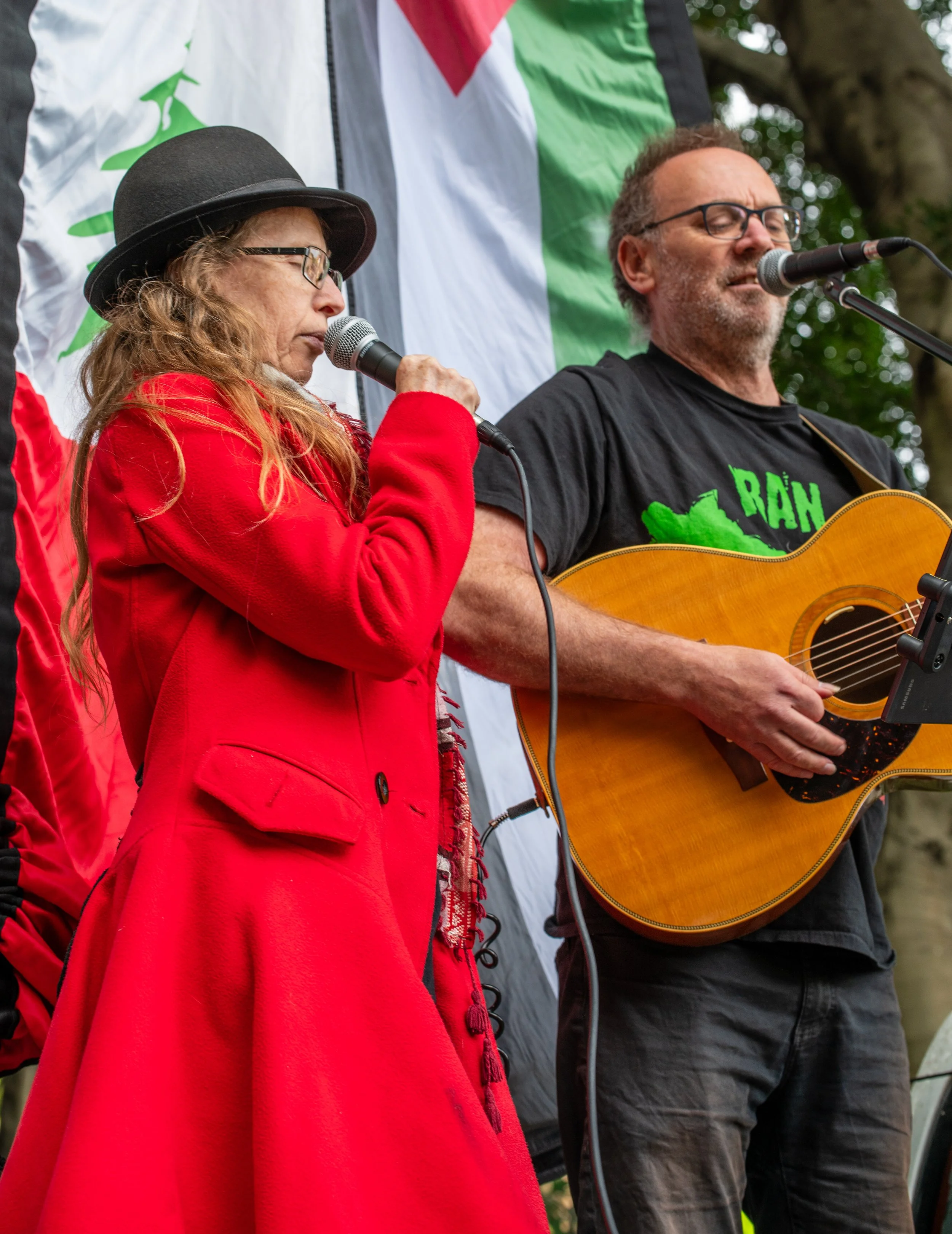 A woman with long red hair, wearing a black hat and red coat, and a man with glasses and a beard, holding an acoustic guitar, singing into microphones during an outdoor event with trees and a flag in the background.
