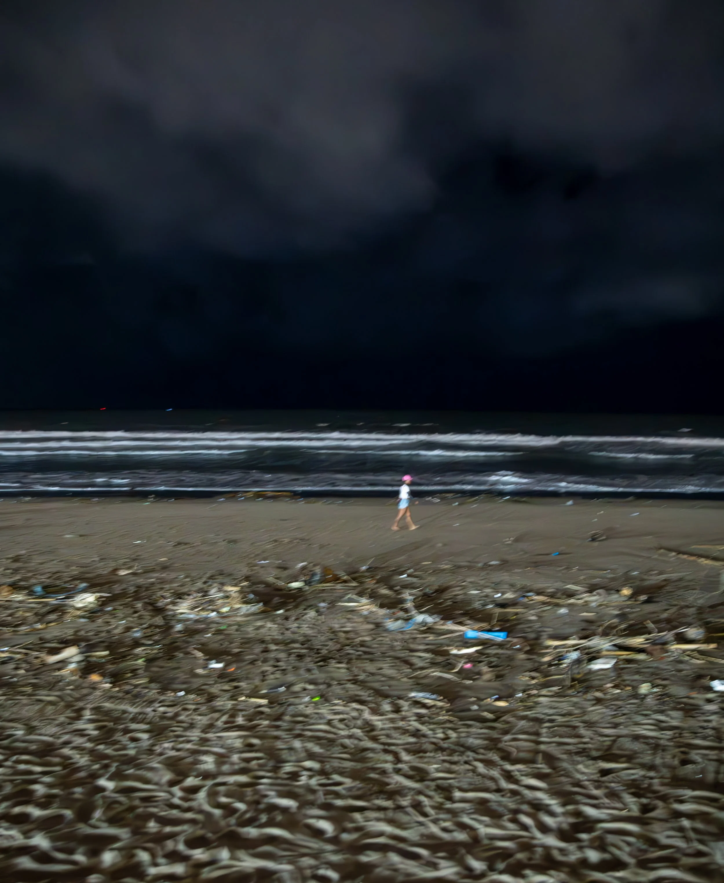A person walking on a beach littered with plastic and trash under a dark, cloudy sky.