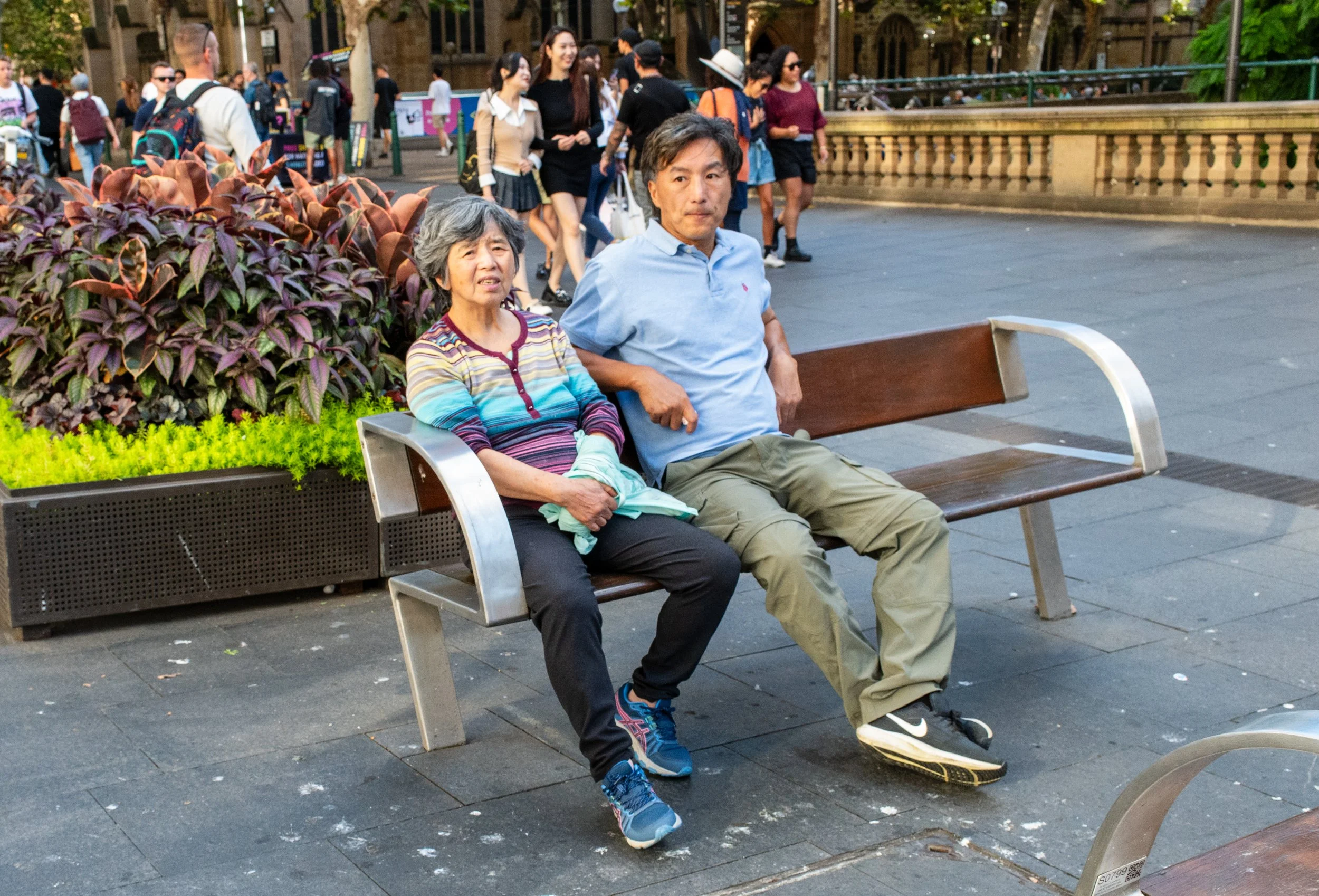 A woman and a man sitting on a park bench surrounded by colorful plants and flowers, with a crowd of pedestrians walking in the background.