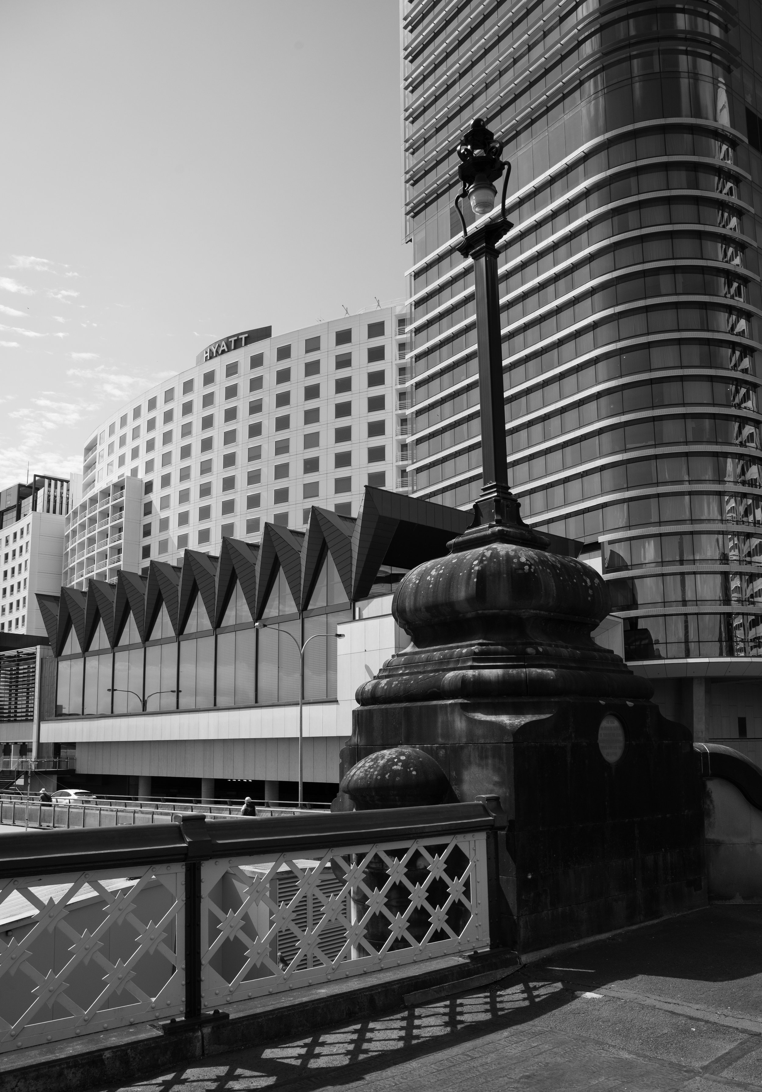 Black and white photo of cityscape with modern buildings, a bridge railing in the foreground, and a city lamp post.