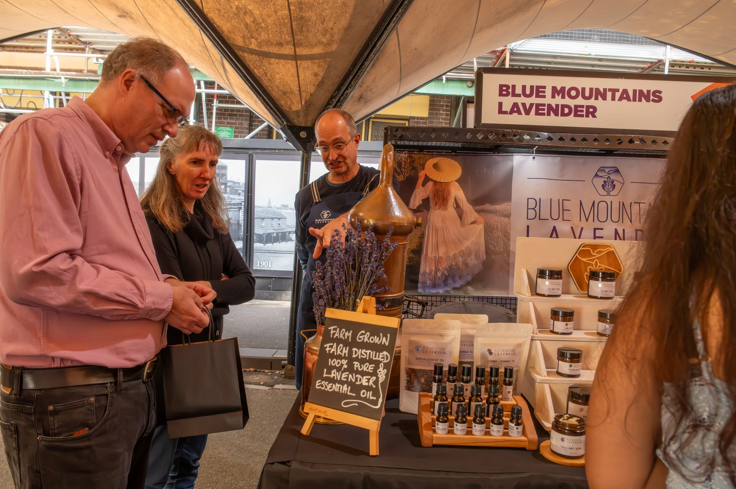 People at a market stall selling lavender products, with a sign reading 'Blue Mountains Lavender', jars, bottles, and lavender bunches on display