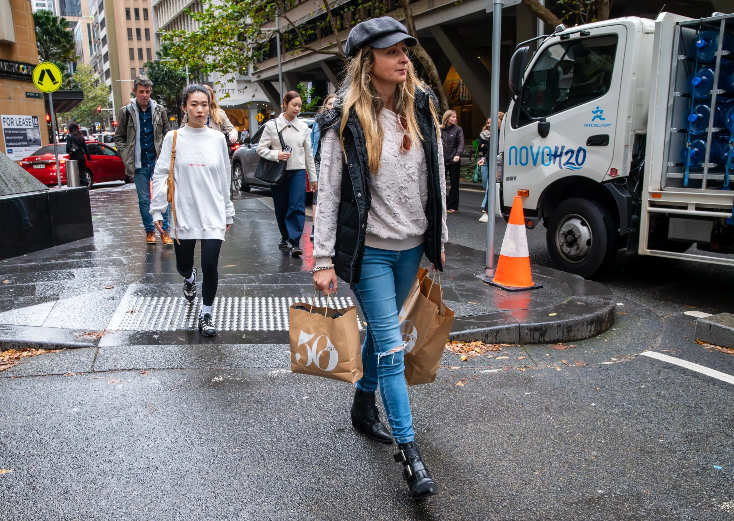People walking on a rainy city sidewalk, carrying shopping bags, with a woman in the foreground wearing a hat, ripped jeans, and a vest, holding shopping bags. There are cars and a delivery truck nearby.