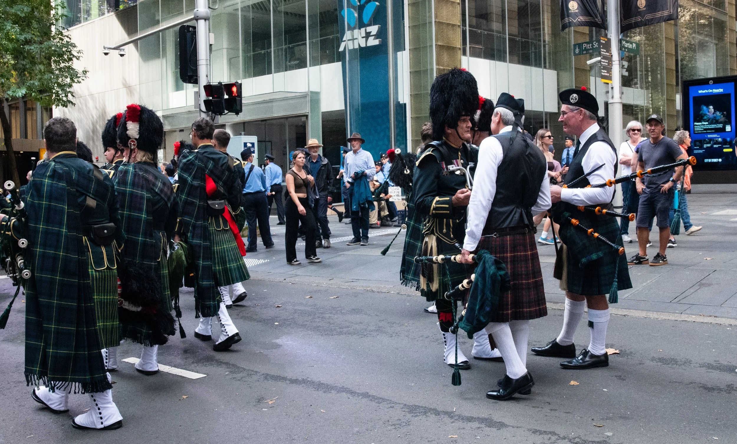 People dressed in traditional Scottish attire, including kilts and tunics, participating in a parade or festival on a city street lined with shops and pedestrians.