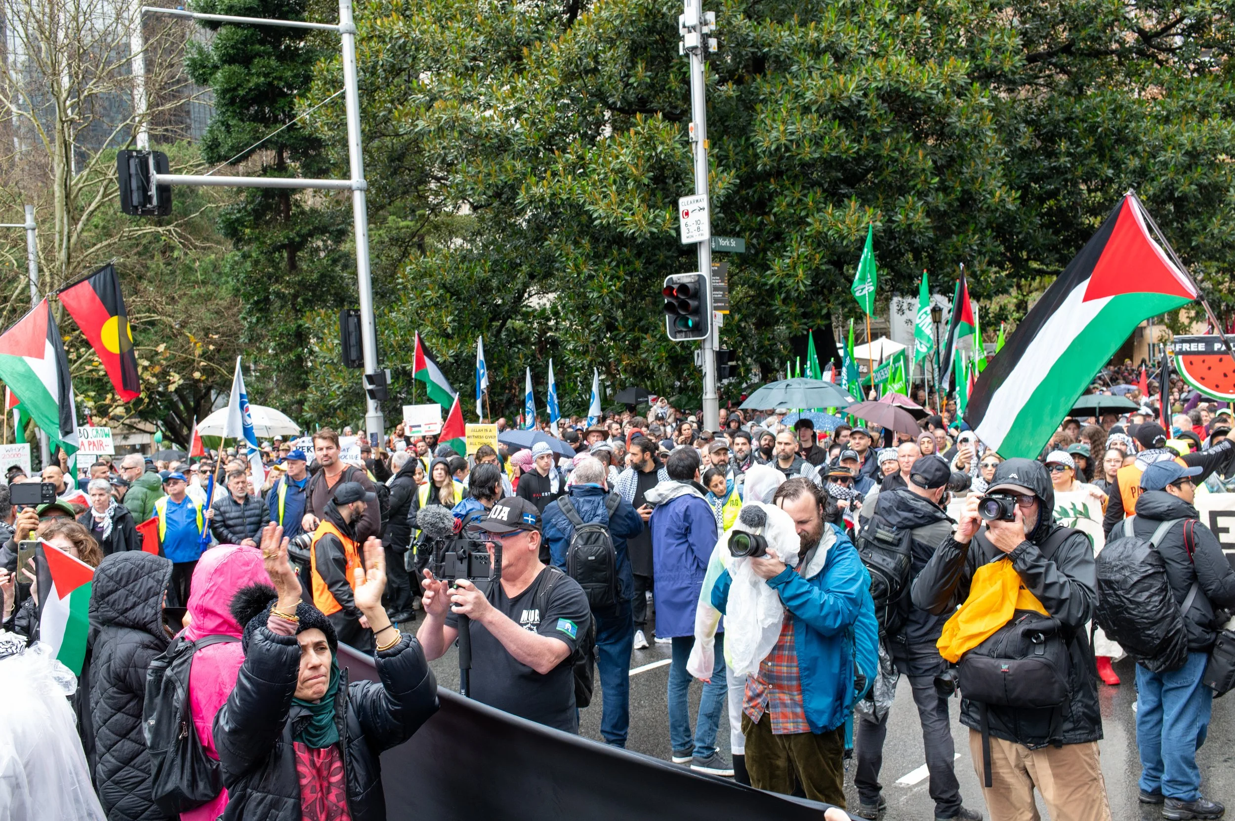 Crowd of protesters with Palestinian flags, some carrying umbrellas, and media personnel with cameras, gathered on a city street during daylight.