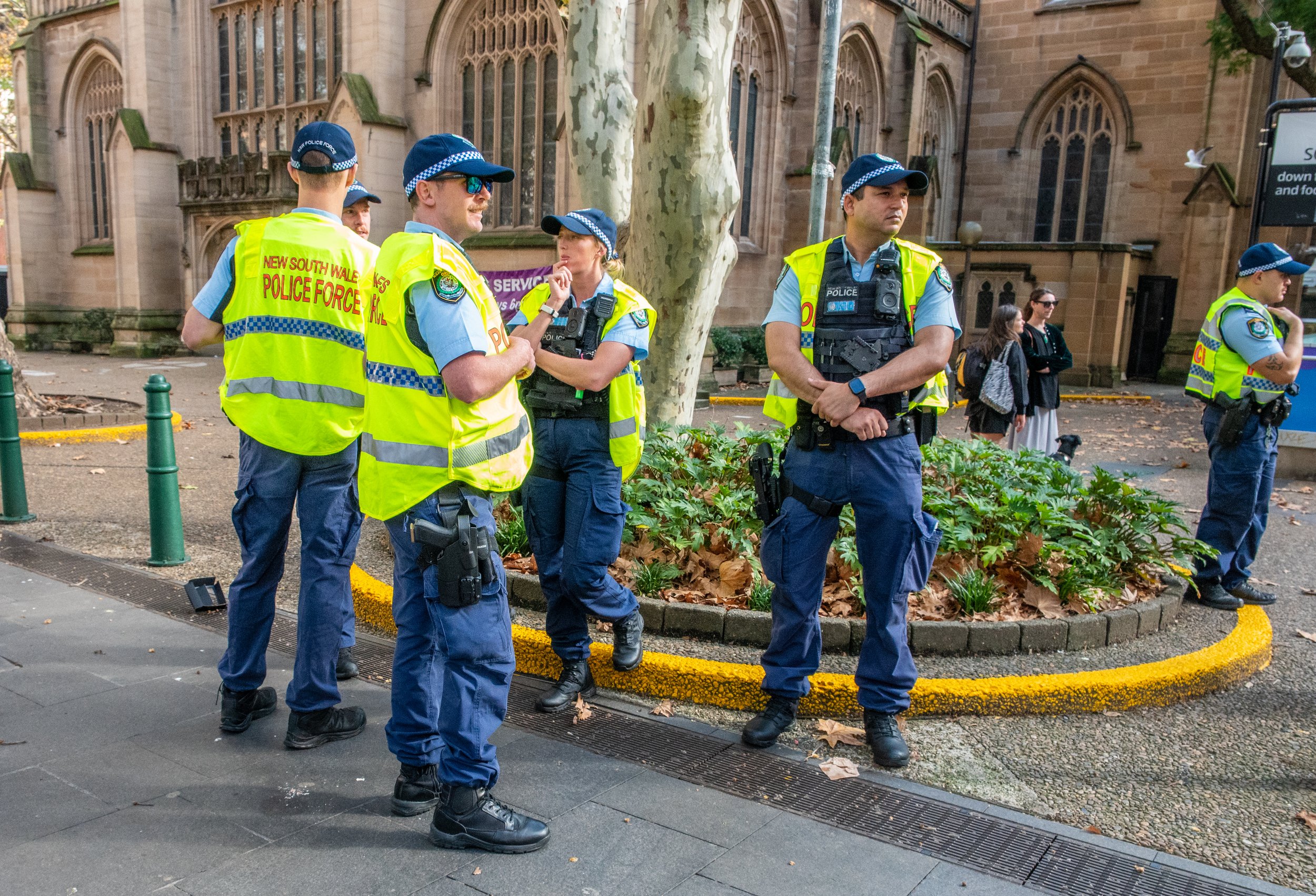 Group of police officers wearing yellow reflective vests and blue uniforms standing and talking outside in front of a historic building with gothic-style windows and a tree with peeling bark.