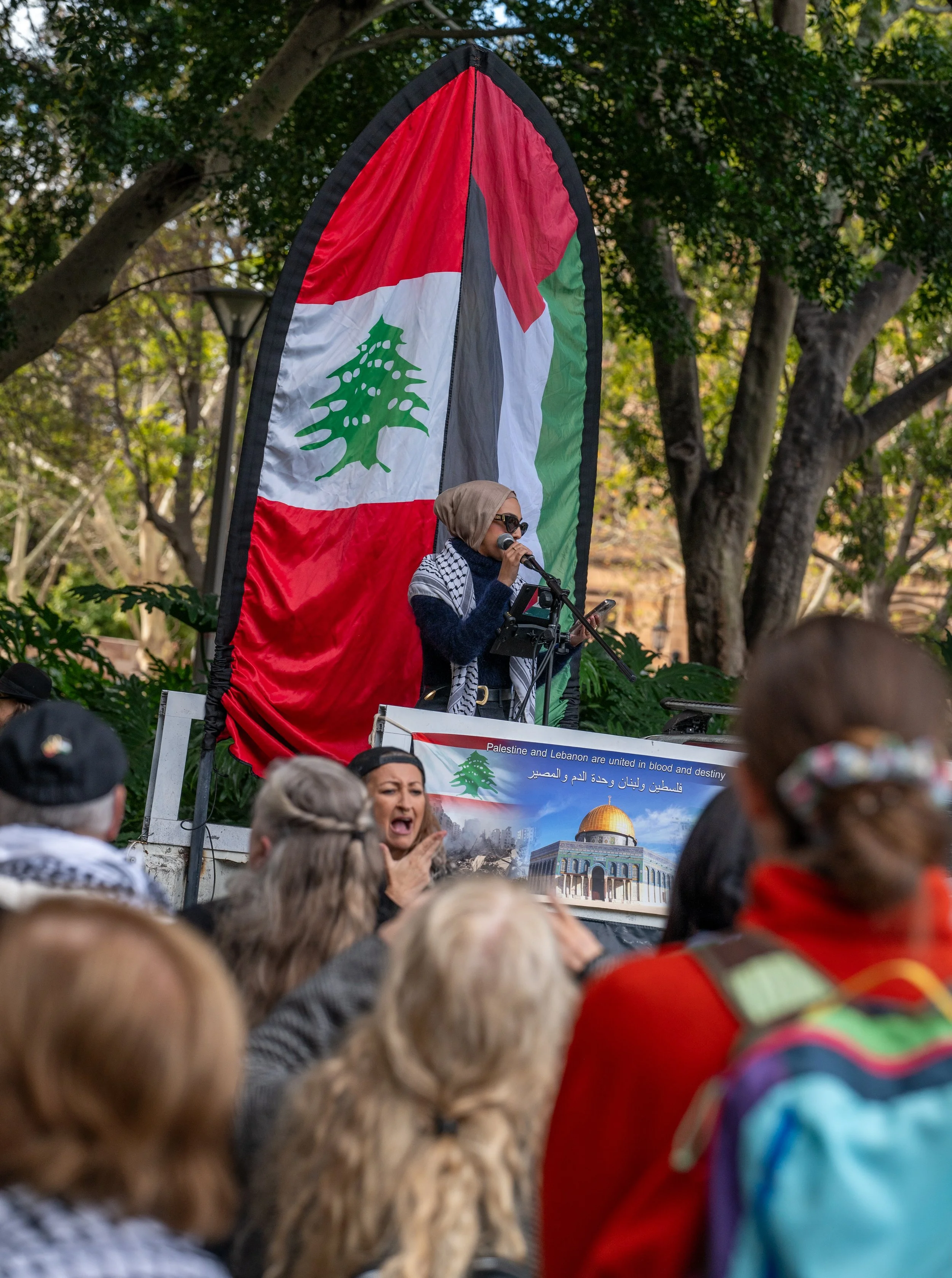 A woman speaking into a microphone at a rally, with a large Lebanese flag as a backdrop, surrounded by a crowd of people.