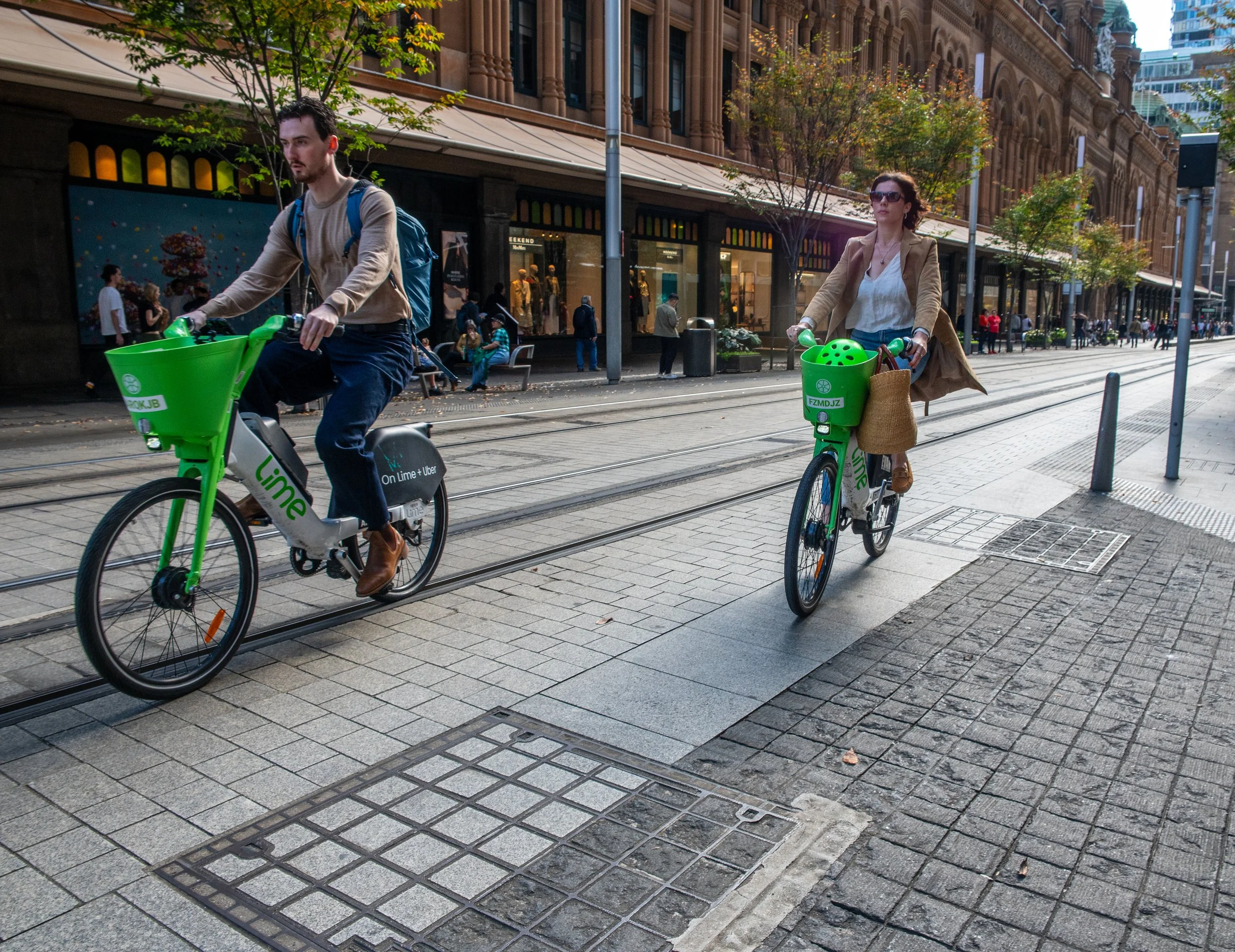 Two people riding Lime electric bikes on city street with tram tracks, sidewalk, and historic buildings in the background.