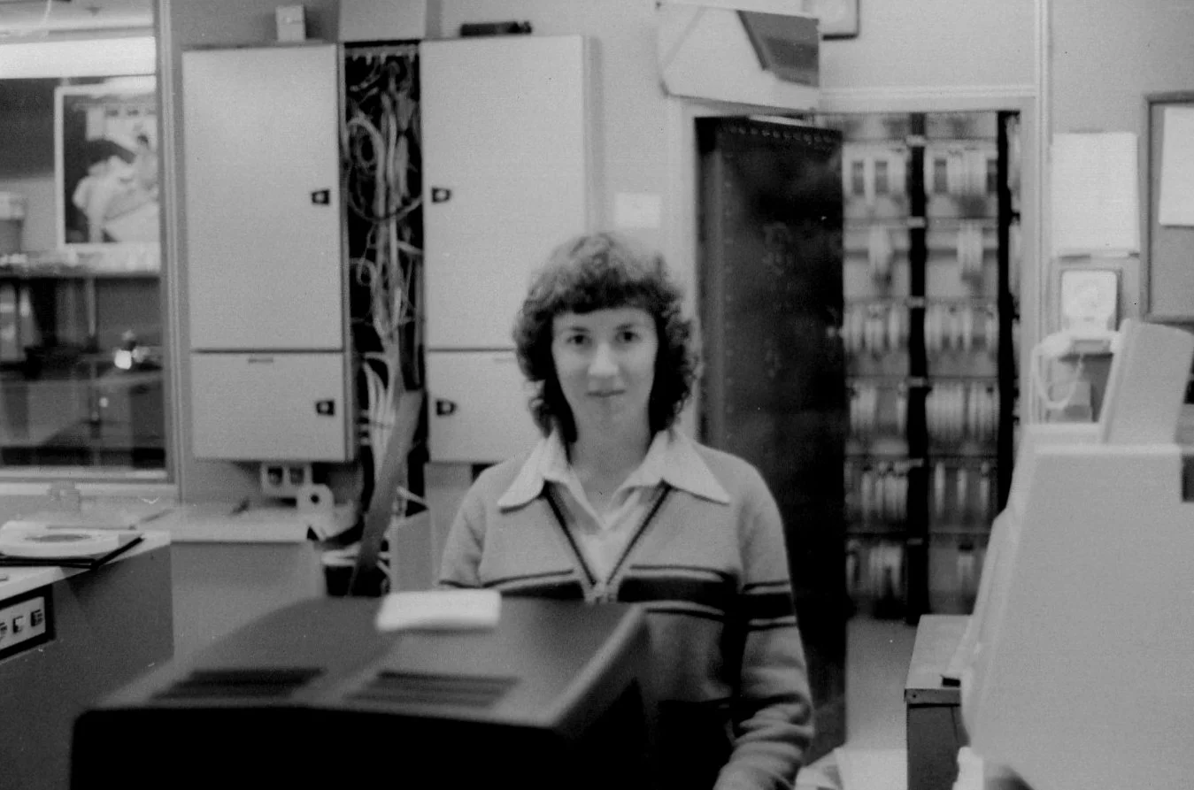A woman with curly hair standing behind a counter in an office or library setting with shelves and electronic equipment in the background.