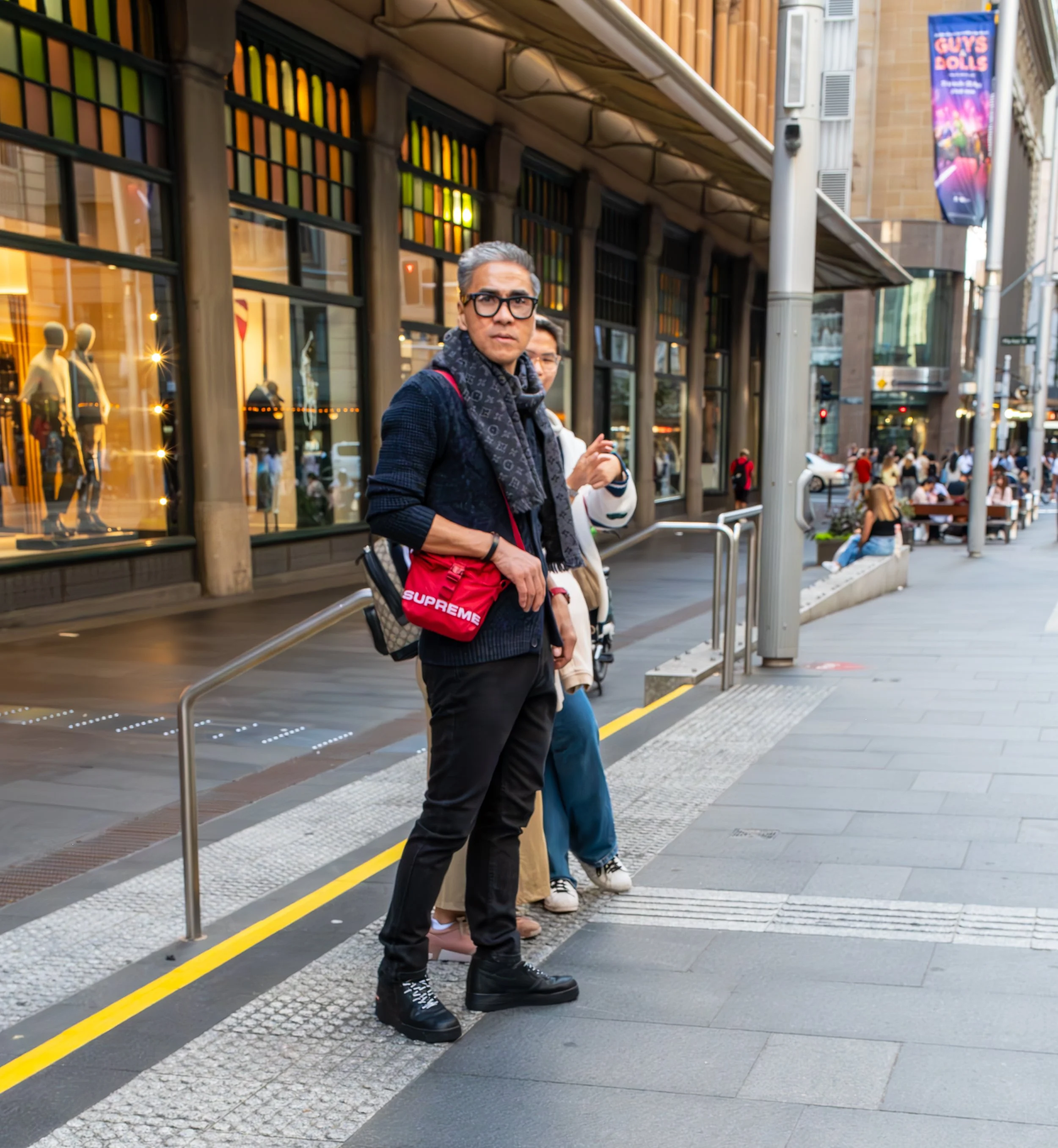 A man with gray hair wearing black glasses, black pants, a dark jacket, and a patterned scarf stands on a city sidewalk with other people nearby. He has a red bag that says "Supreme" across his chest, and he is looking directly at the camera. The background shows a building with large windows and a busy street with pedestrians and outdoor seating.