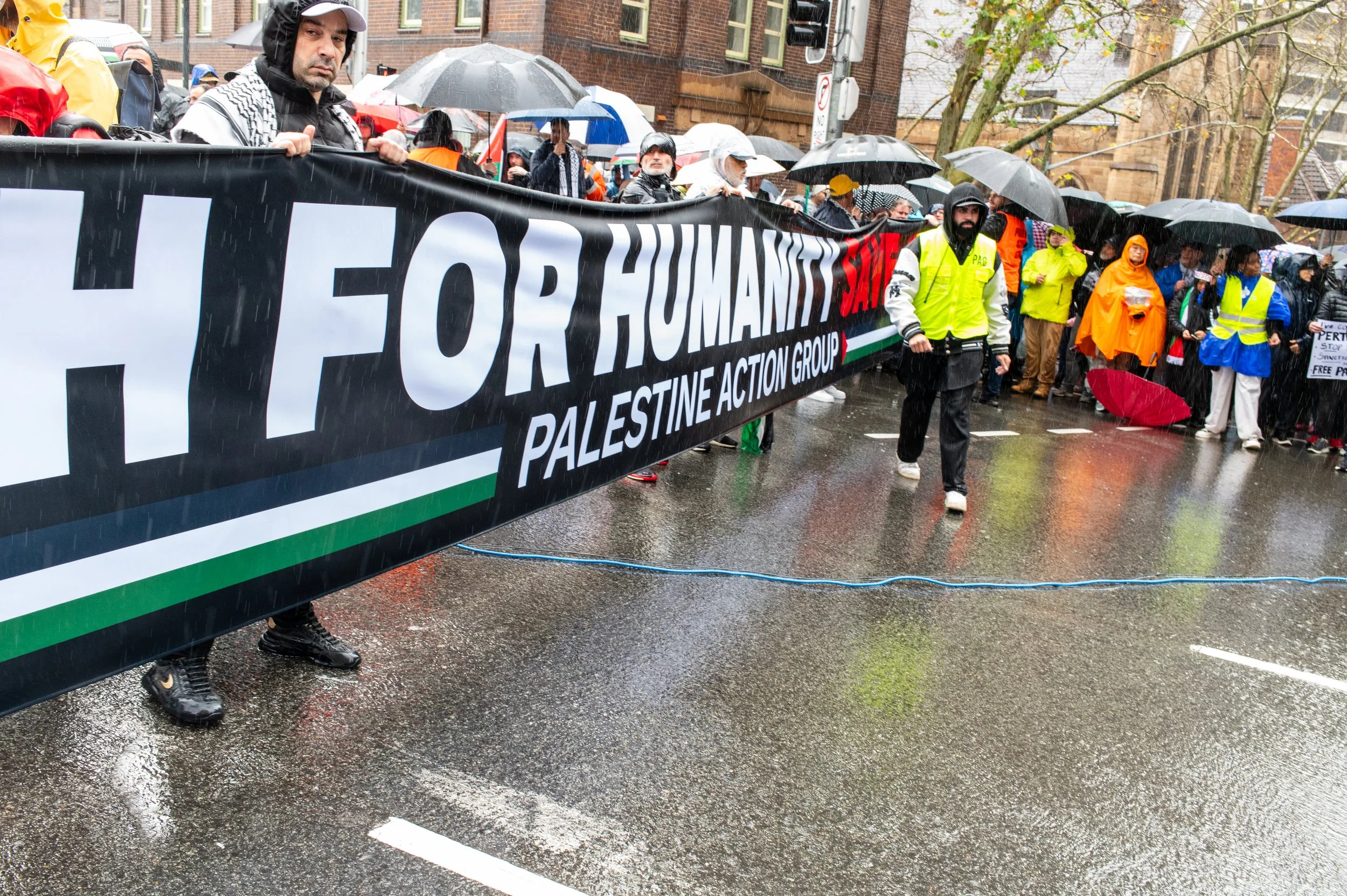 Protesters holding a large banner that reads 'H for Humanity, Palestine Action Group' during a rainy demonstration on a city street with many people, umbrellas, and wet pavement.