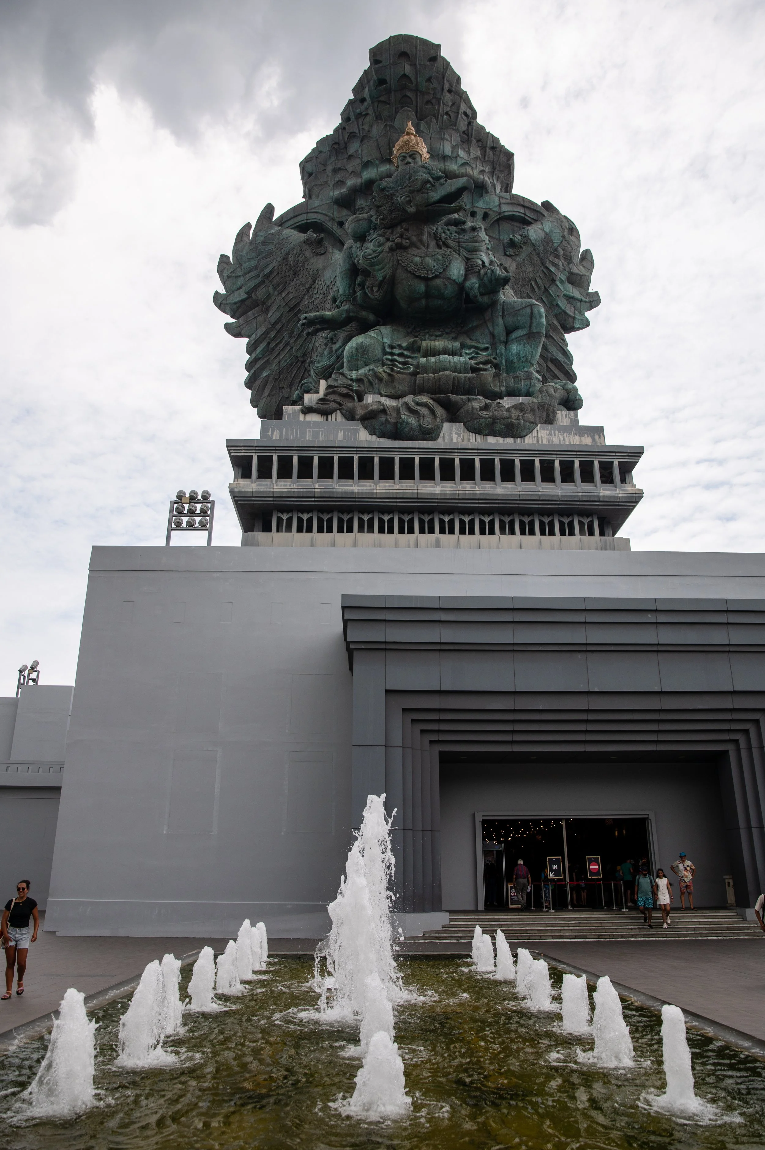 A large, dark bronze statue of Garuda, a mythical bird, situated outside a modern building with a fountain in the foreground and cloudy sky overhead.