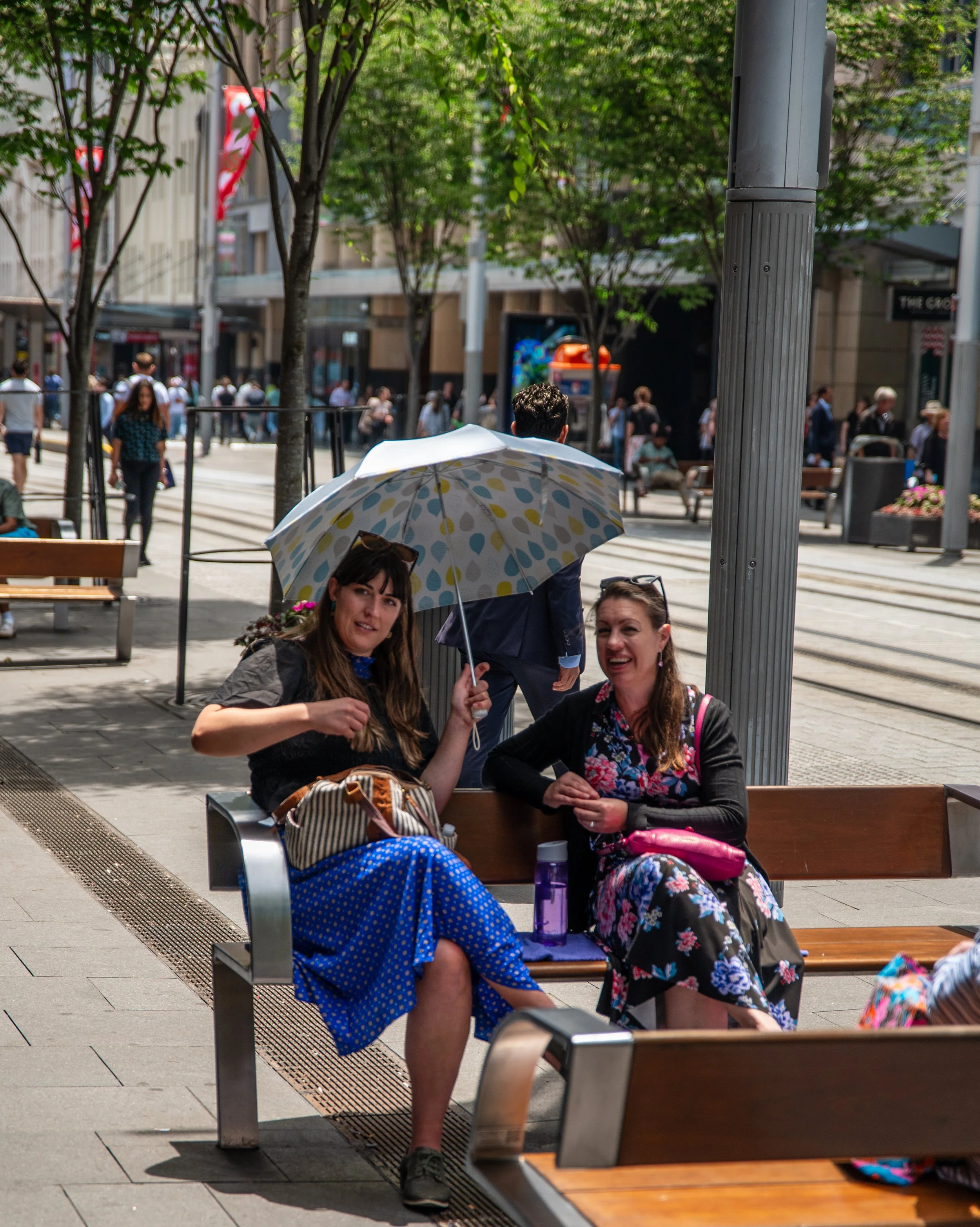 Two women sitting on a park bench under an umbrella, smiling and talking, with a busy city street in the background.