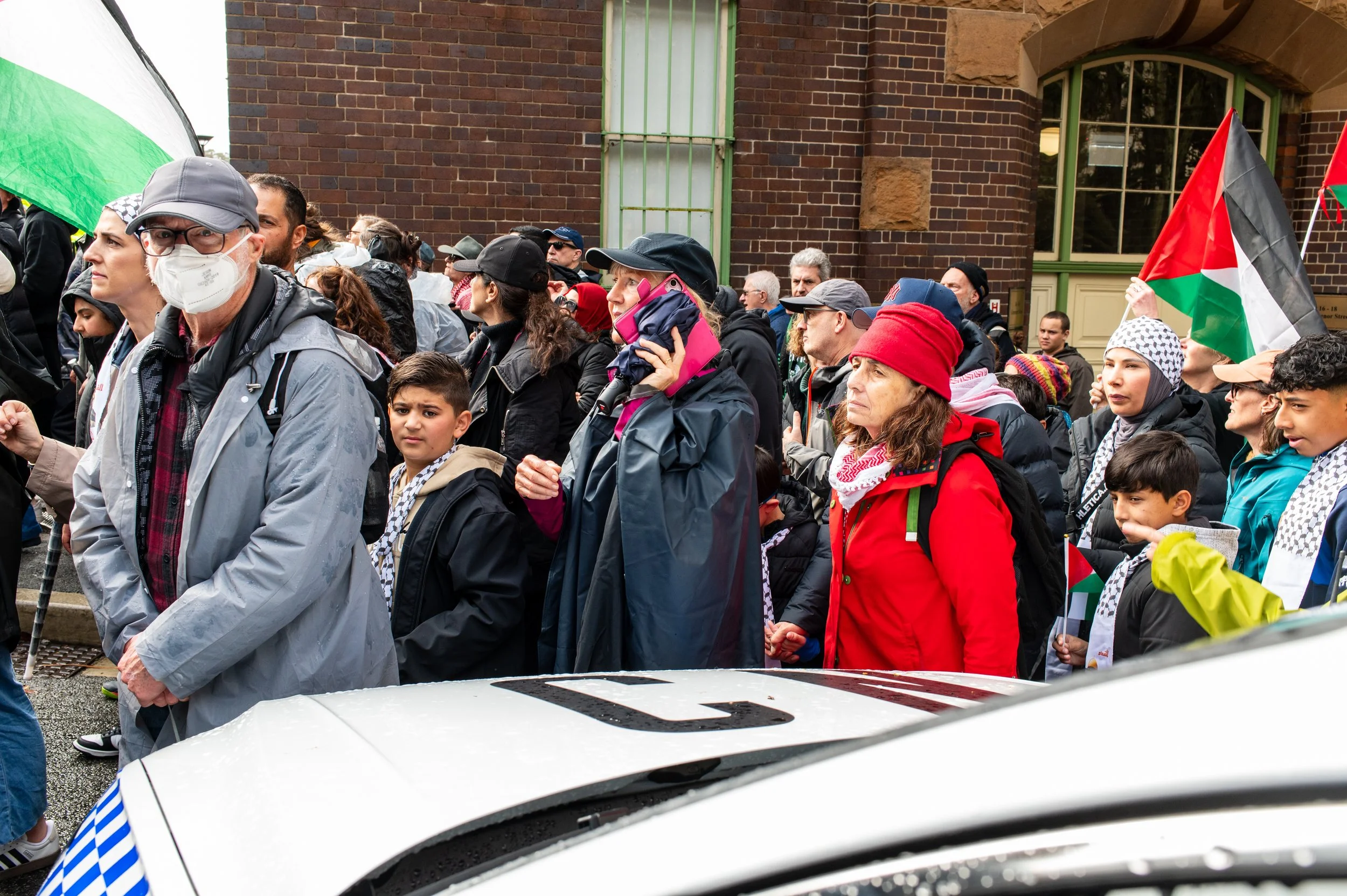 A large group of diverse people gathered in a protest or demonstration, some holding Palestinian flags. Many are wearing jackets, hats, and scarves, with one woman talking on her cellphone. A police car is visible in the foreground.