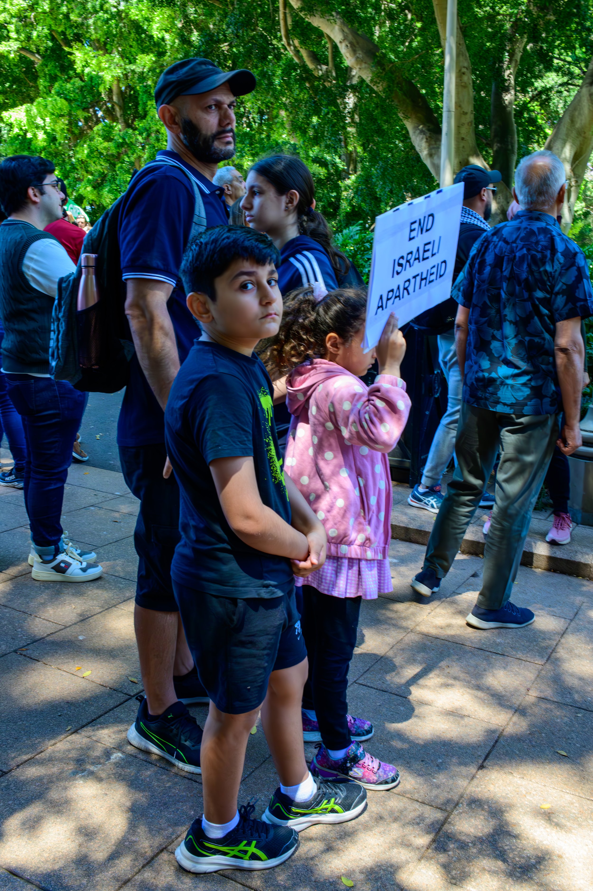 A diverse group of people protesting, holding a sign that reads 'END ISRAELI APARTHEID' in a park with trees.