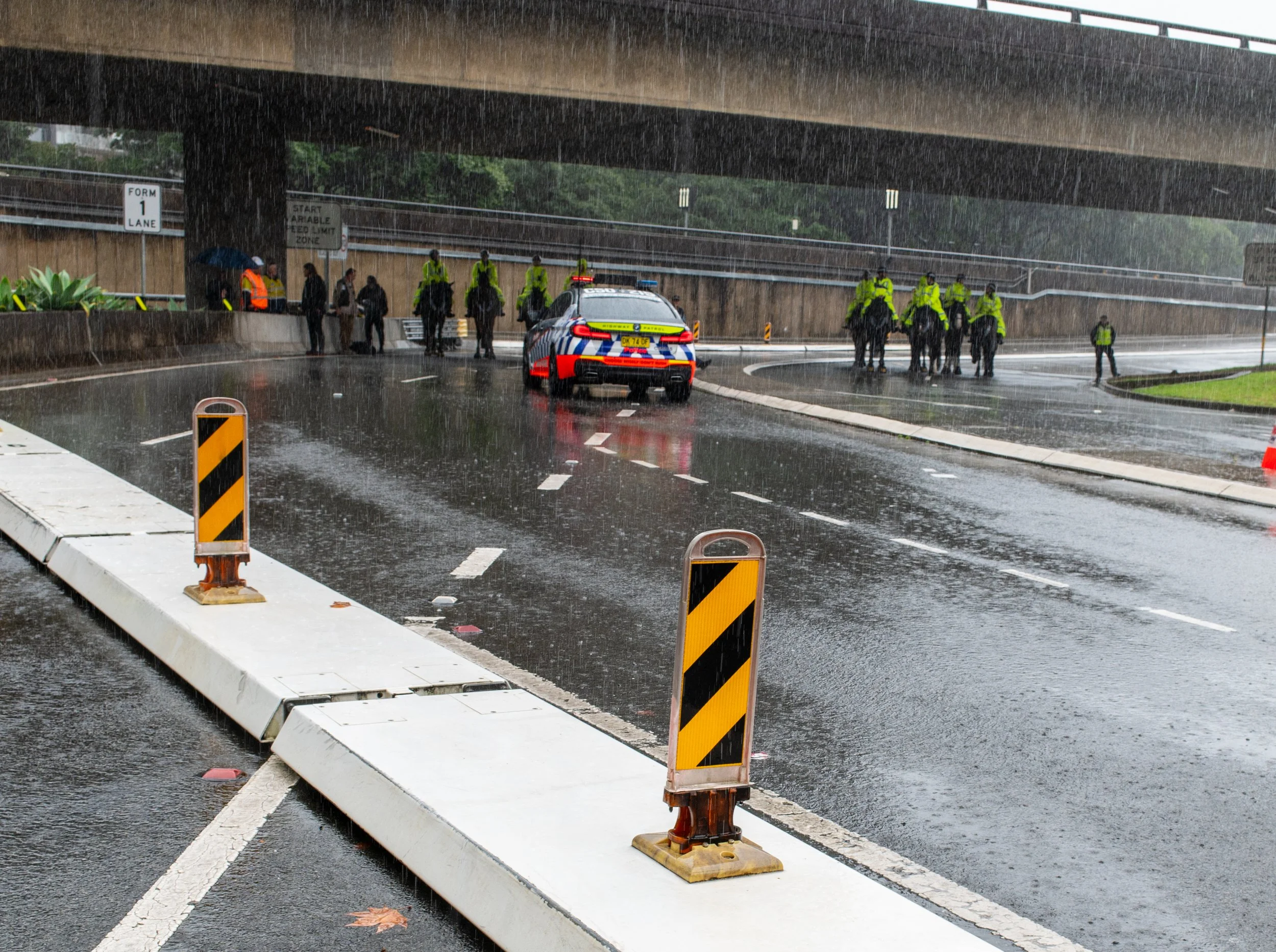 Police officers and police car under a bridge on a rainy day, with traffic barricades on wet road.