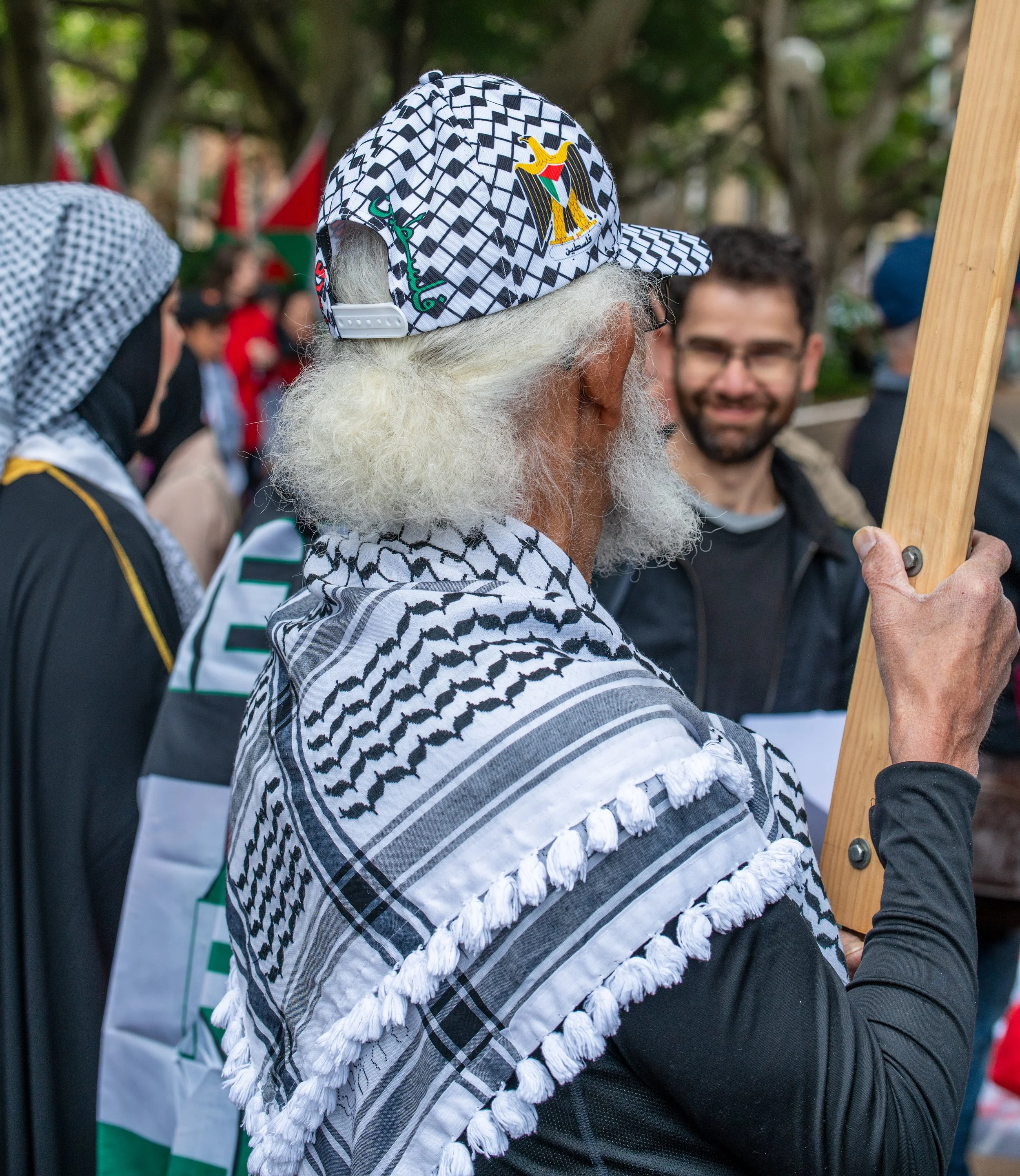 An elderly man with white hair and beard wearing a Palestinian keffiyeh and a cap with Palestinian symbols, holding a wooden stick during a gathering.