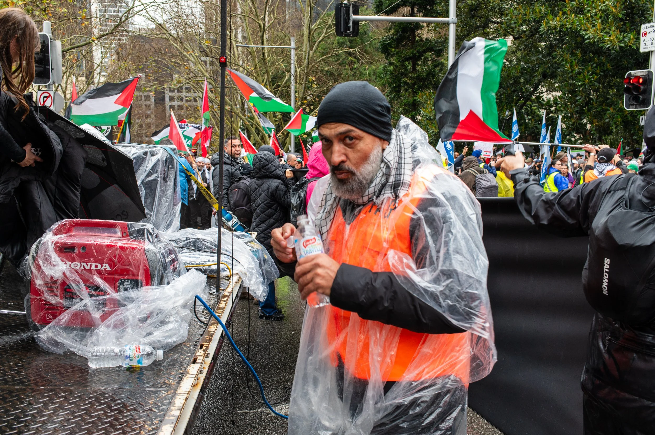 A man wearing a black head covering, orange safety vest, and a plastic rain poncho stands in front of a crowd with Palestinian flags during a rainy day protest. He is holding a water bottle, and there is a table with a red portable generator and water bottles in front of him. Many people in the crowd are dressed warmly, and trees line the background.