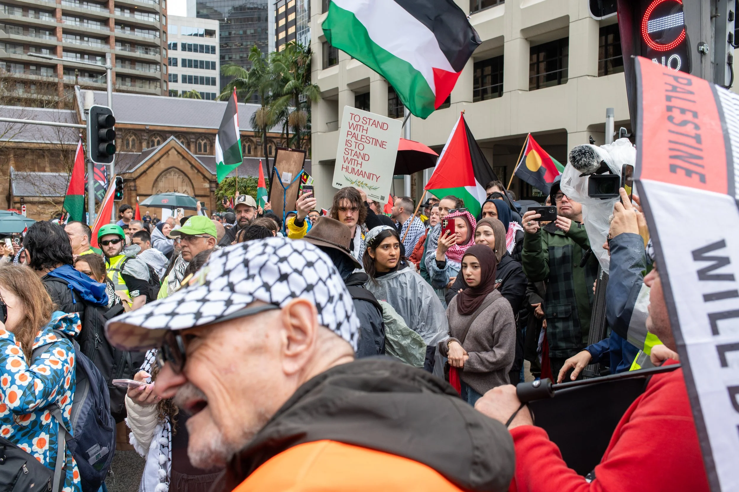 Crowd of protesters with Palestinian flags, signs, and umbrellas during a demonstration, with buildings and palm trees in the background.