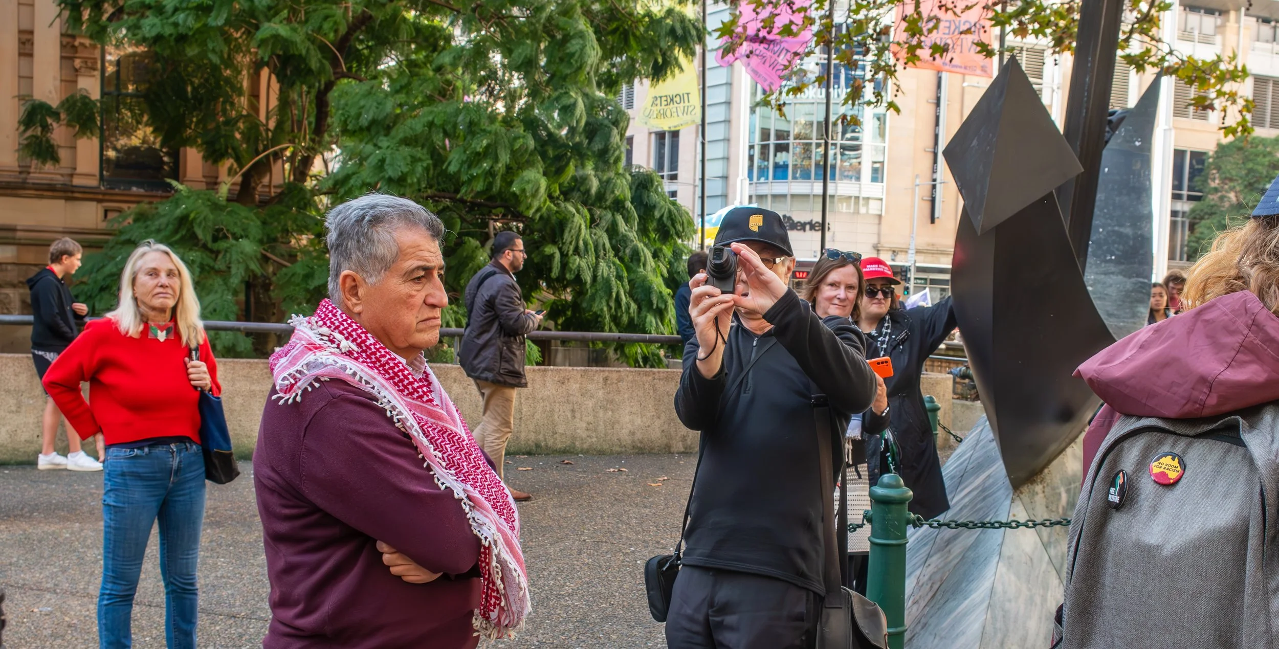 People gathered outdoors near a monument or sculpture, with some taking photos or observing, during daytime. The scene includes a man in a purple jacket and red scarf, a woman in a red sweater, and others in casual attire. There are trees, buildings, and banners in the background.