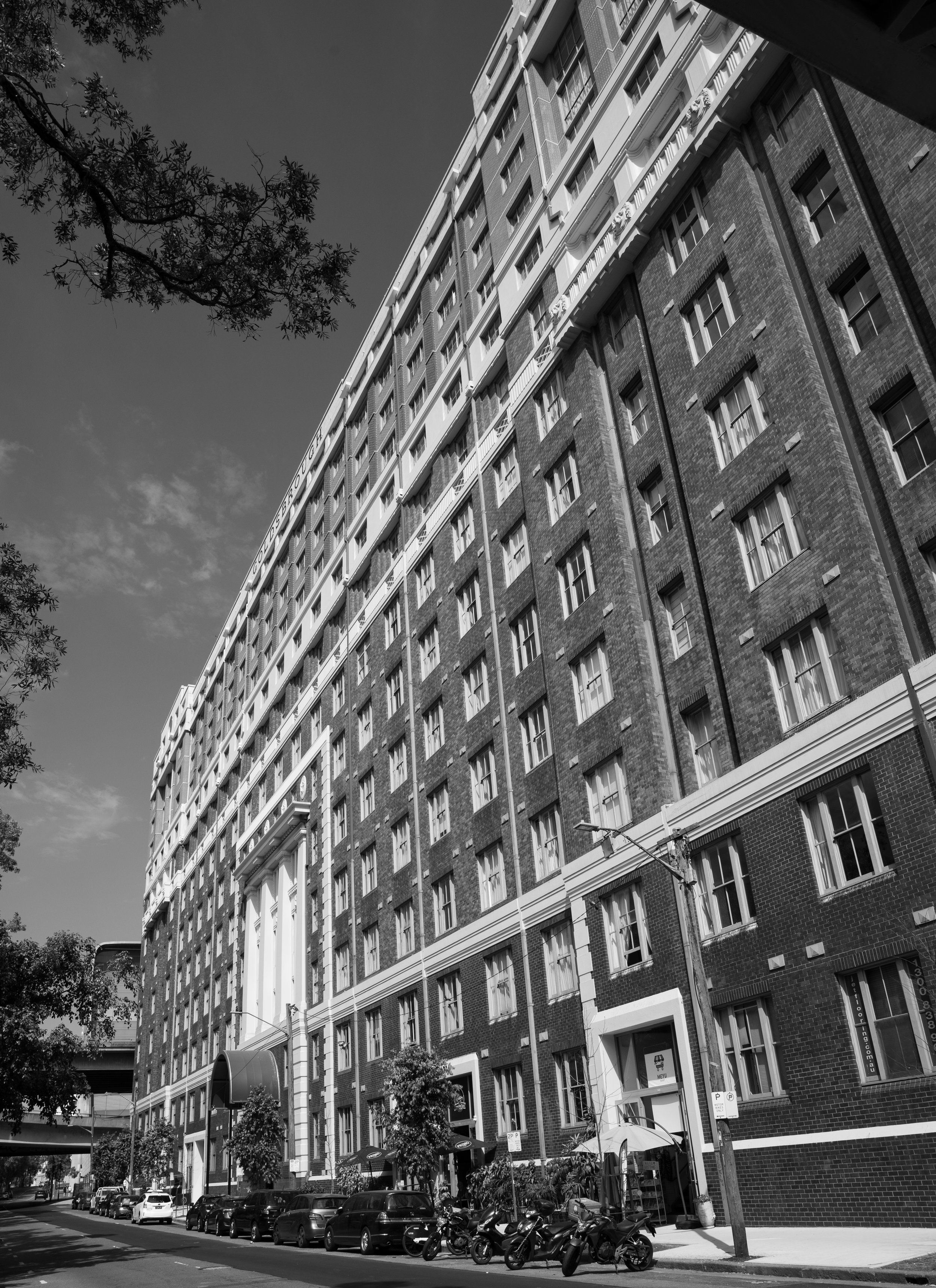 A tall multi-story brick apartment building with numerous windows, seen from the street level in black and white.