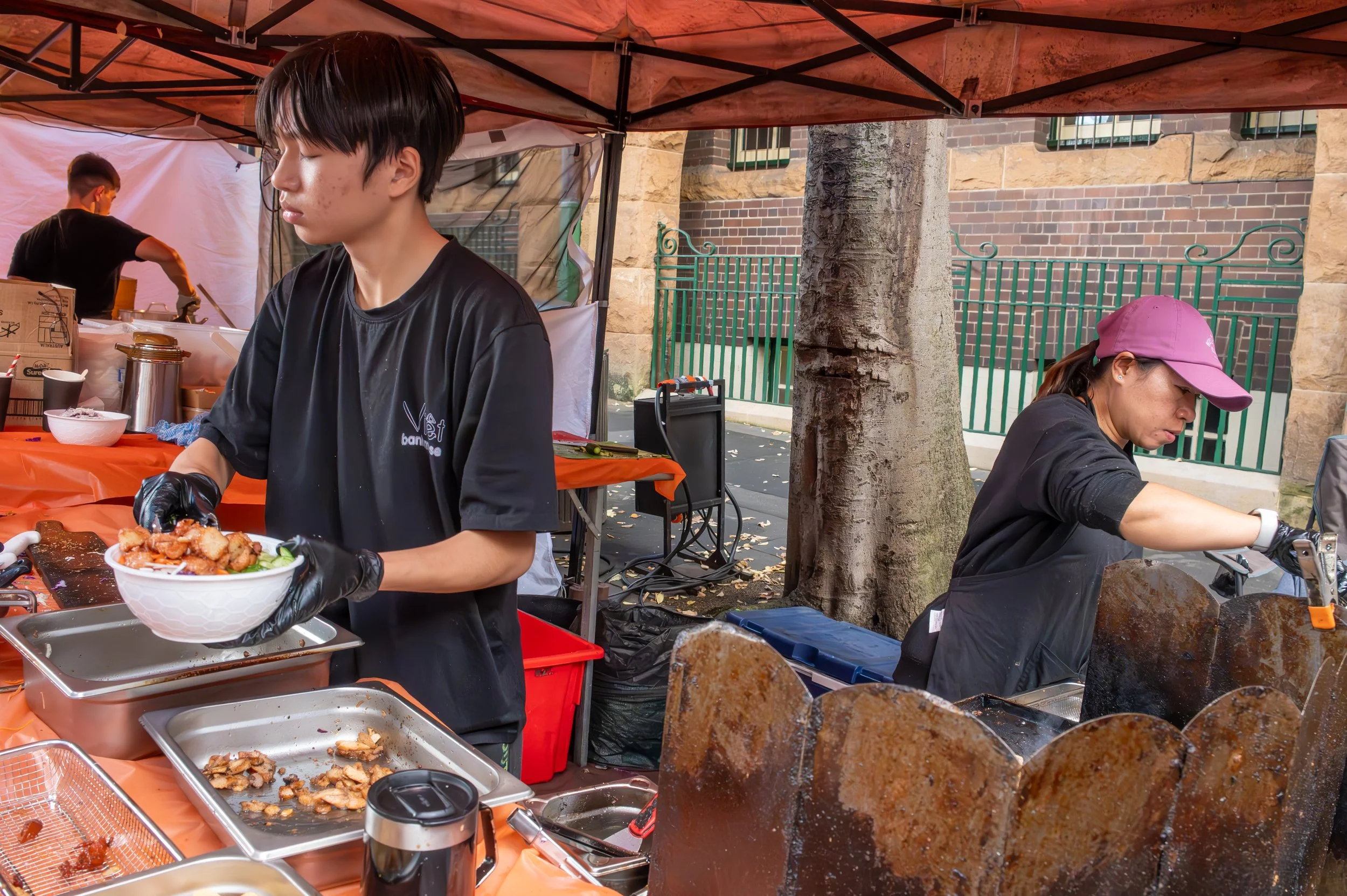 Two women working at a food stall outdoors, one is serving food into a bowl, and the other is grilling meat on a large metal grill, with a tree and green fence in the background.