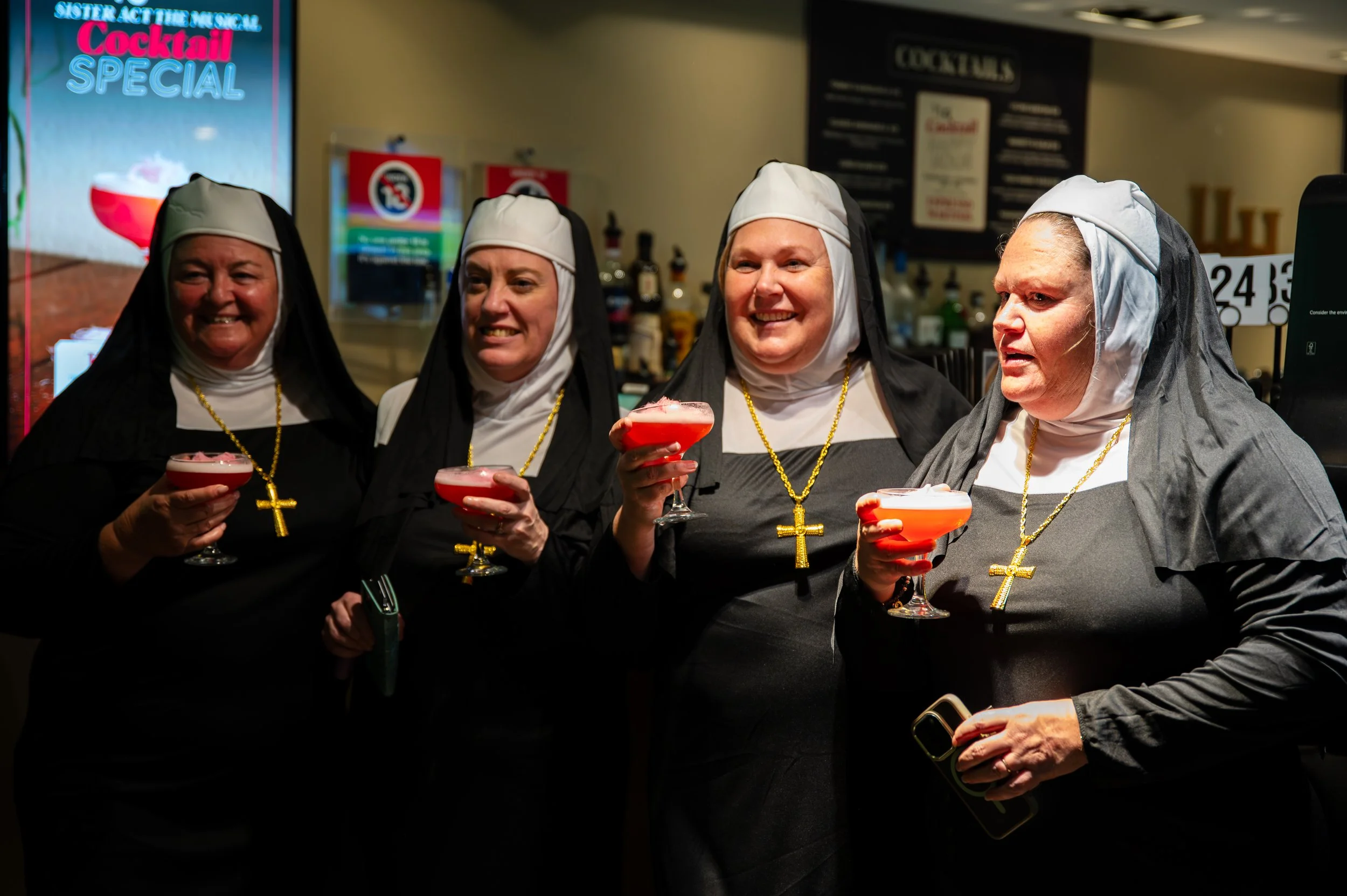 Four women dressed as nuns holding pink cocktails at a bar or restaurant, smiling and posing for a photo.