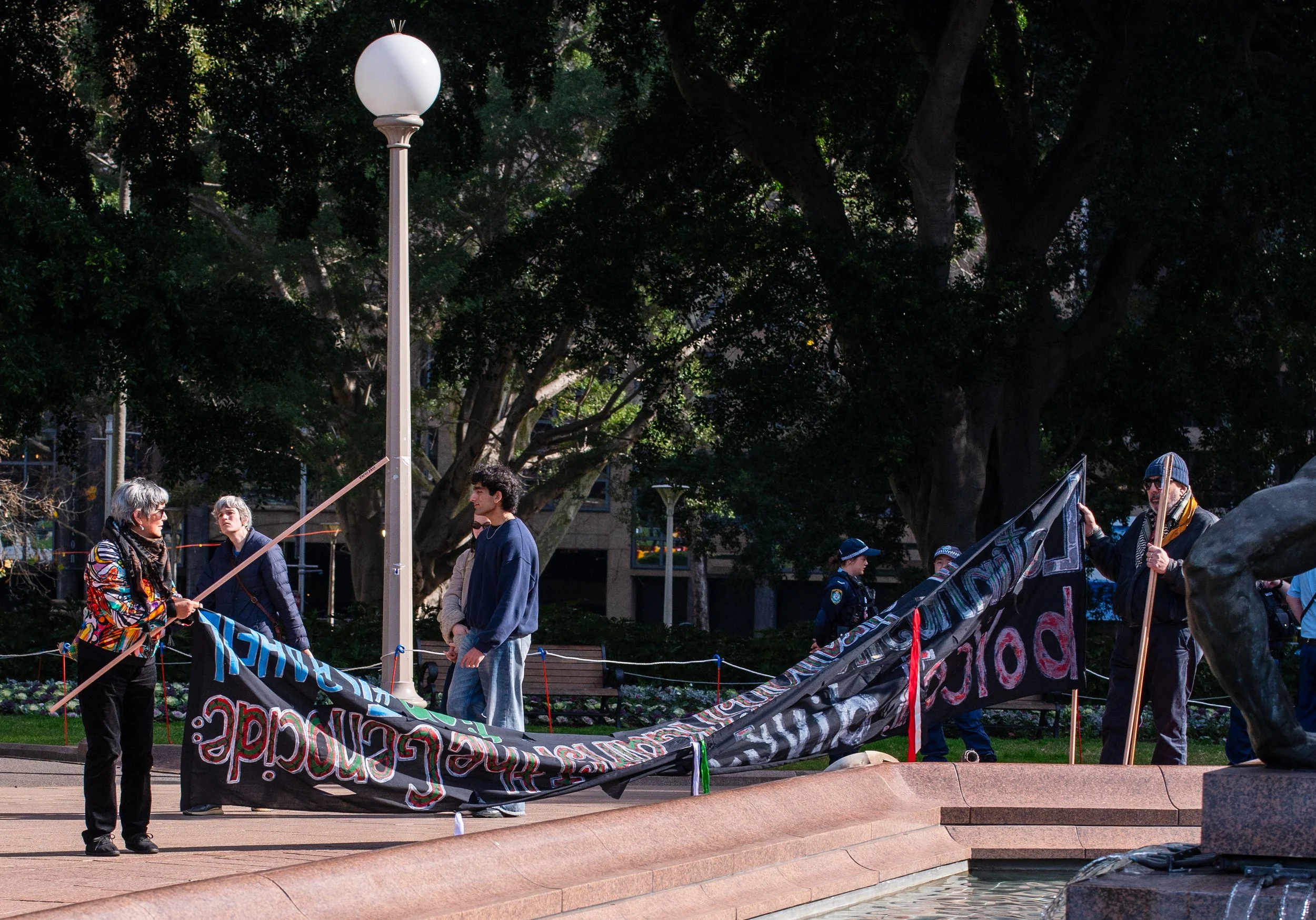 People holding a large banner and a flag in a park during a protest or demonstration, with police officers observing in the background.