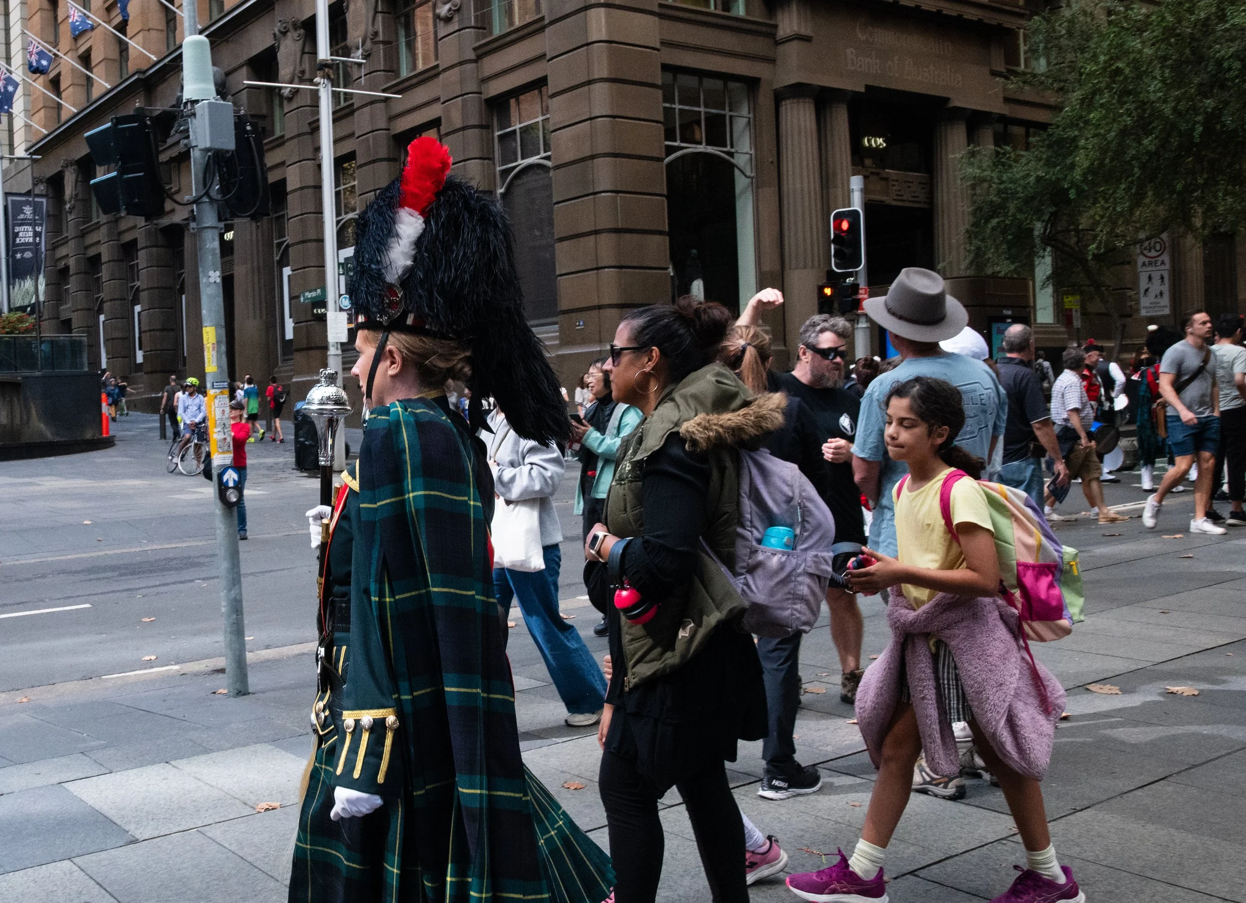 Crowd of people walking across a city intersection, with a person in a Scottish military uniform and a tall hat in the foreground.